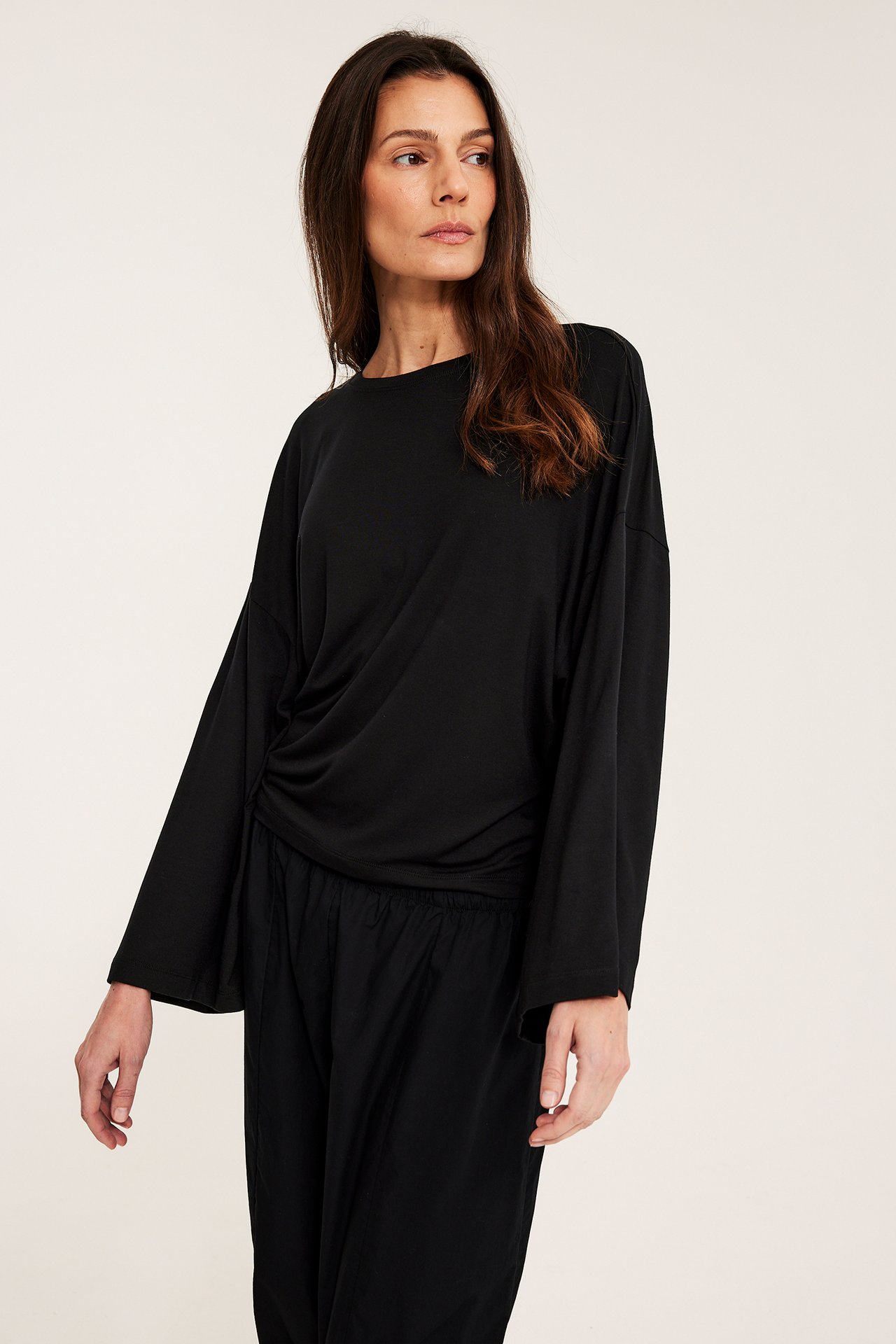 Framsida av kvinna i en svart, draperad långärmad topp med vida ärmar och svarta byxor.