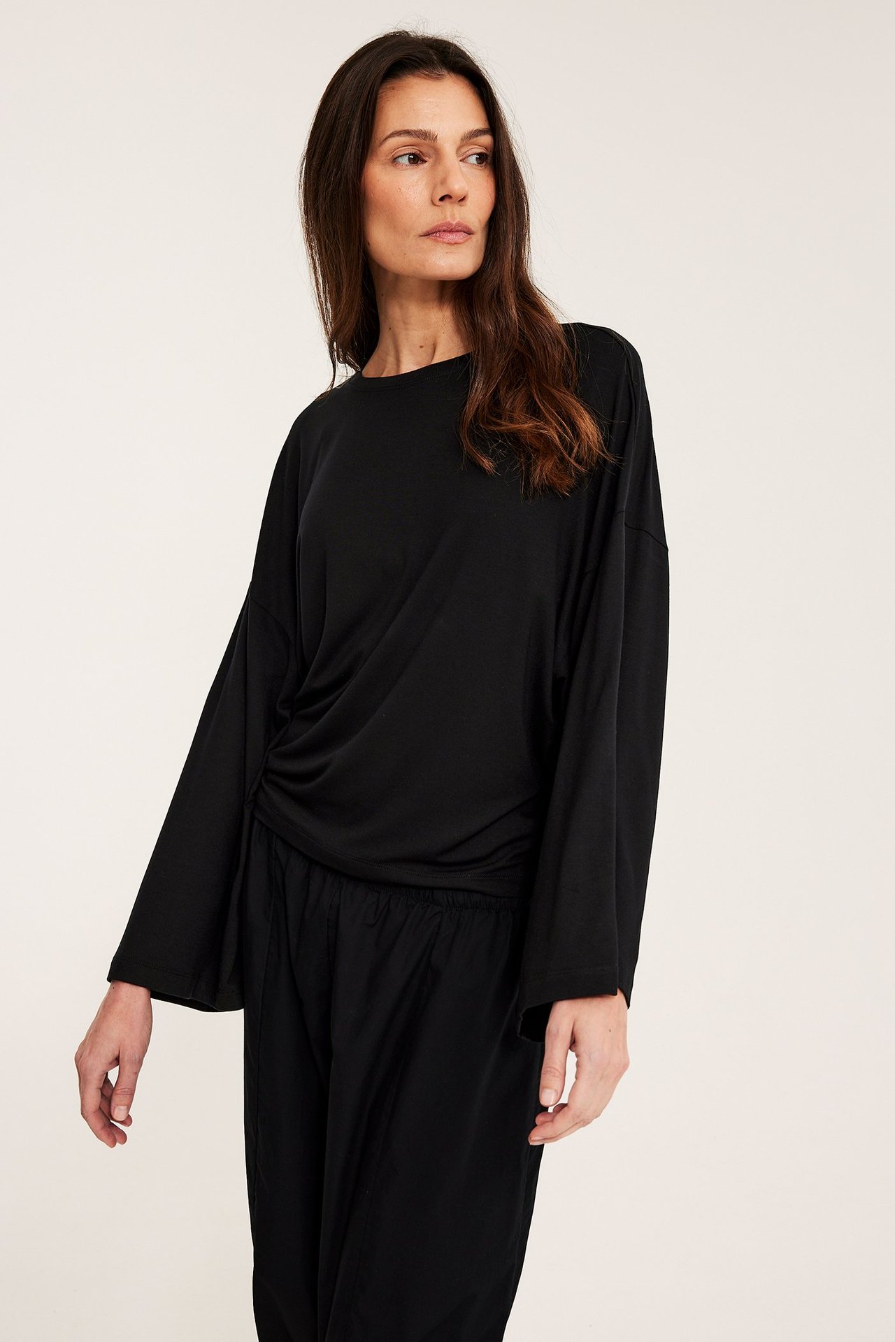 Widok z przodu kobiety w czarnym, lejącym topie z długimi i szerokimi rękawami oraz czarnych spodniach.