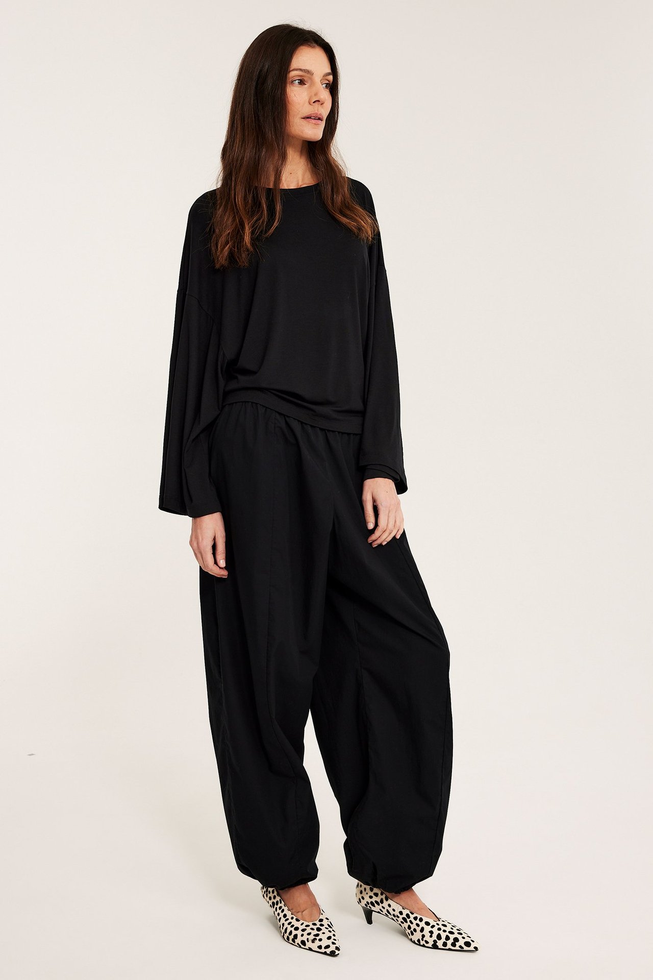 Widok z boku kobiety w czarnej bluzce z szerokimi rękawami, czarnych spodniach i szpilkach w panterkę.