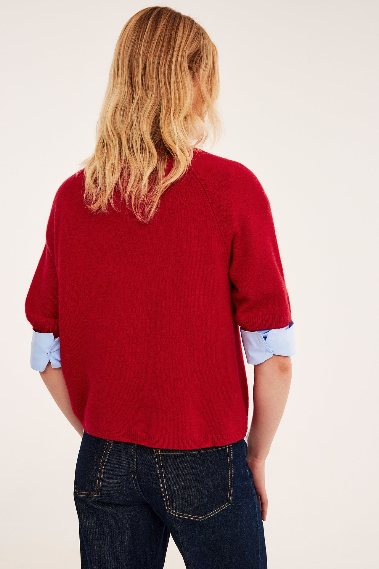 Kvinne bakfra, rød kortermet strikkegenser over blå skjorte, mørke jeans.