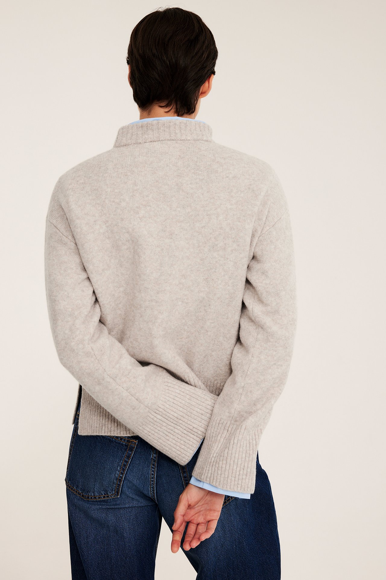 Ryggvy: kvinna i beige melerad stickad tröja över ljusblå skjorta och mörkblå jeans.