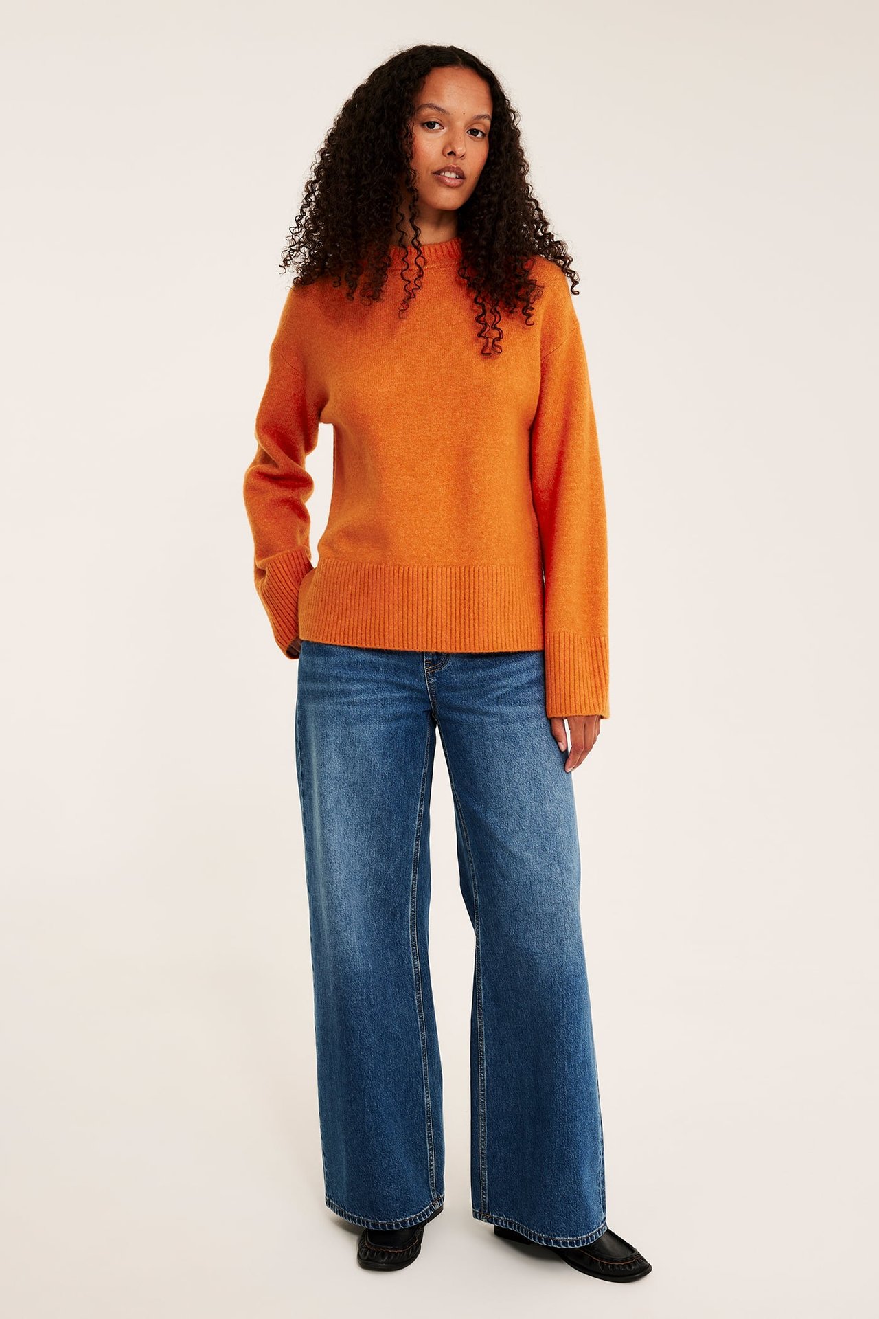 Frontbilde av en kvinne i oransje rundhalset strikkegenser, blå vide jeans og svarte sko.
