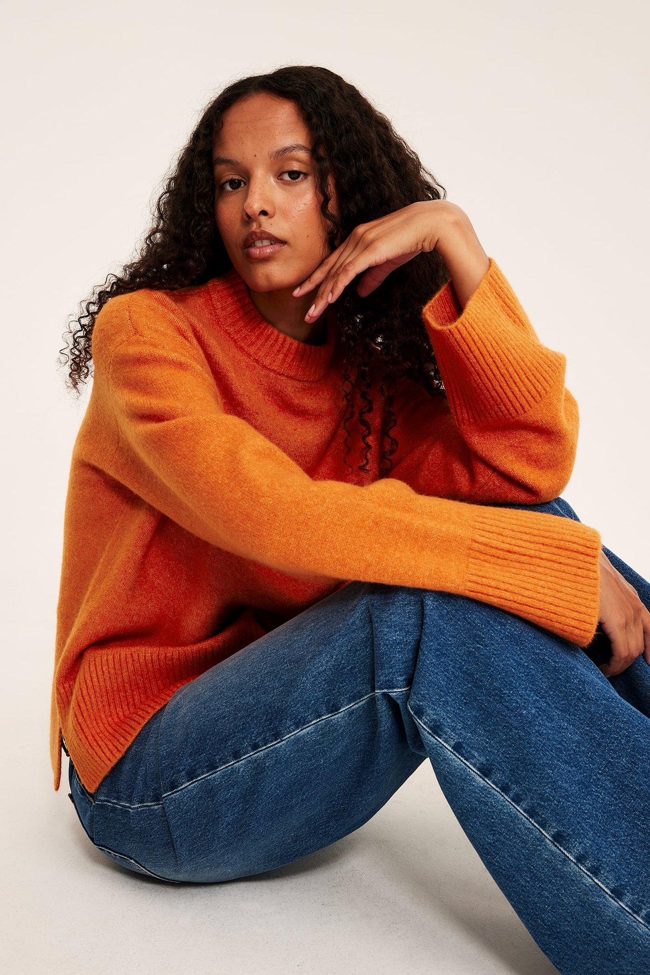 Frontbilde av kvinnelig modell i oransje rundhalset genser og blå jeans.