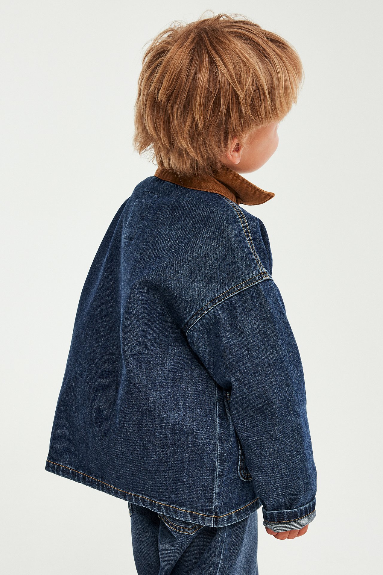 Bakifrån: Barn i mörkblå lös jeansjacka med brun manchesterkrage och matchande jeans.