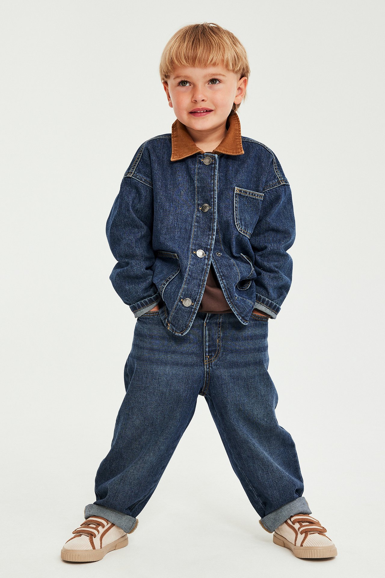 Barn i mörkblå jeansjacka med brun krage, matchande jeans och beiga sneakers, framifrån.