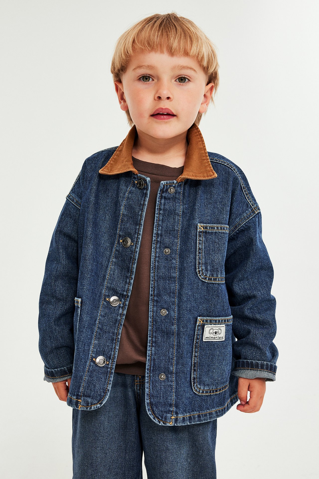 Front view: Barn i blå jeansjacka med brun krage, brun t-shirt och jeansbyxor.