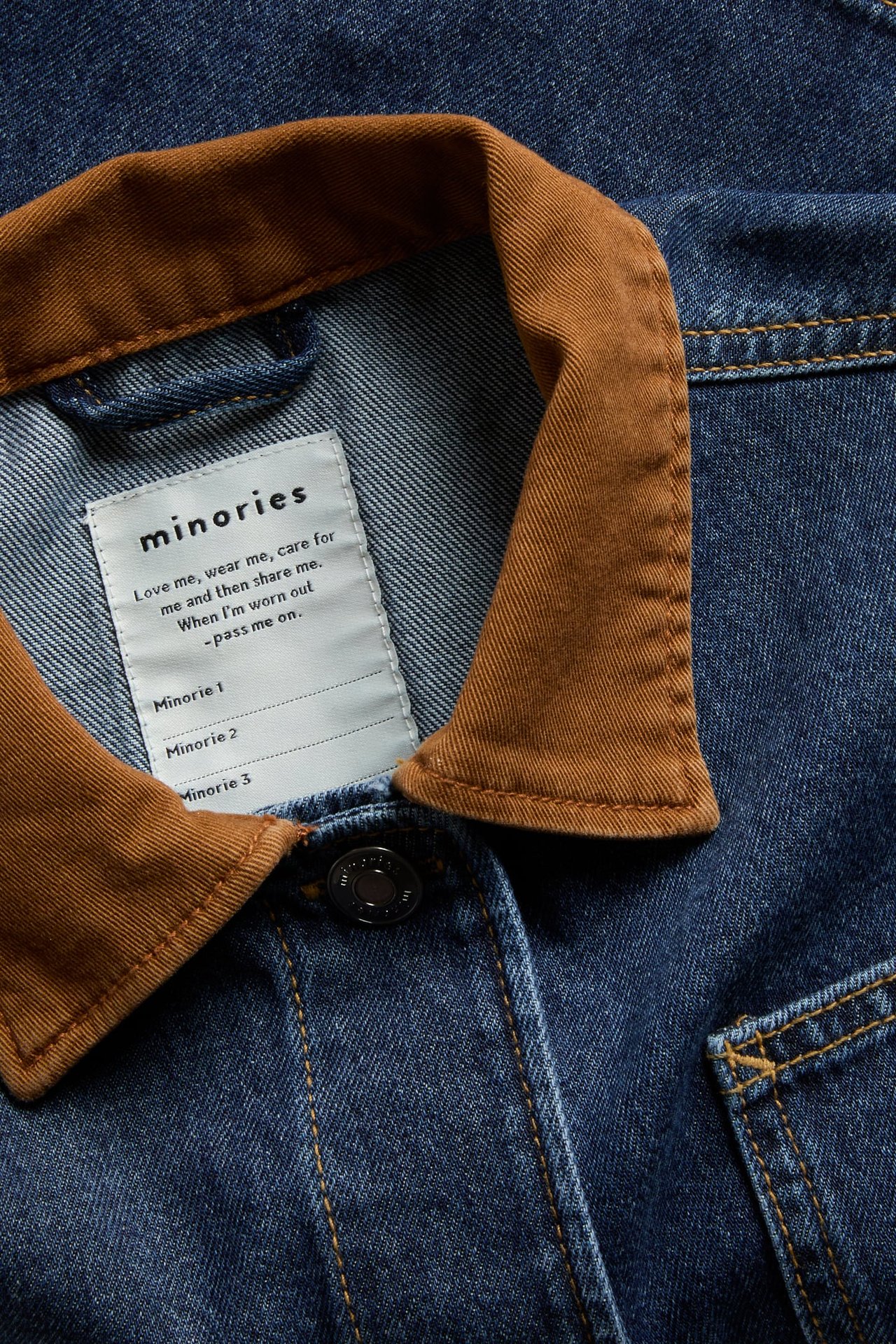 Zbliżenie niebieskiej kurtki jeansowej z brązowym kołnierzem i białą metką „minories”.