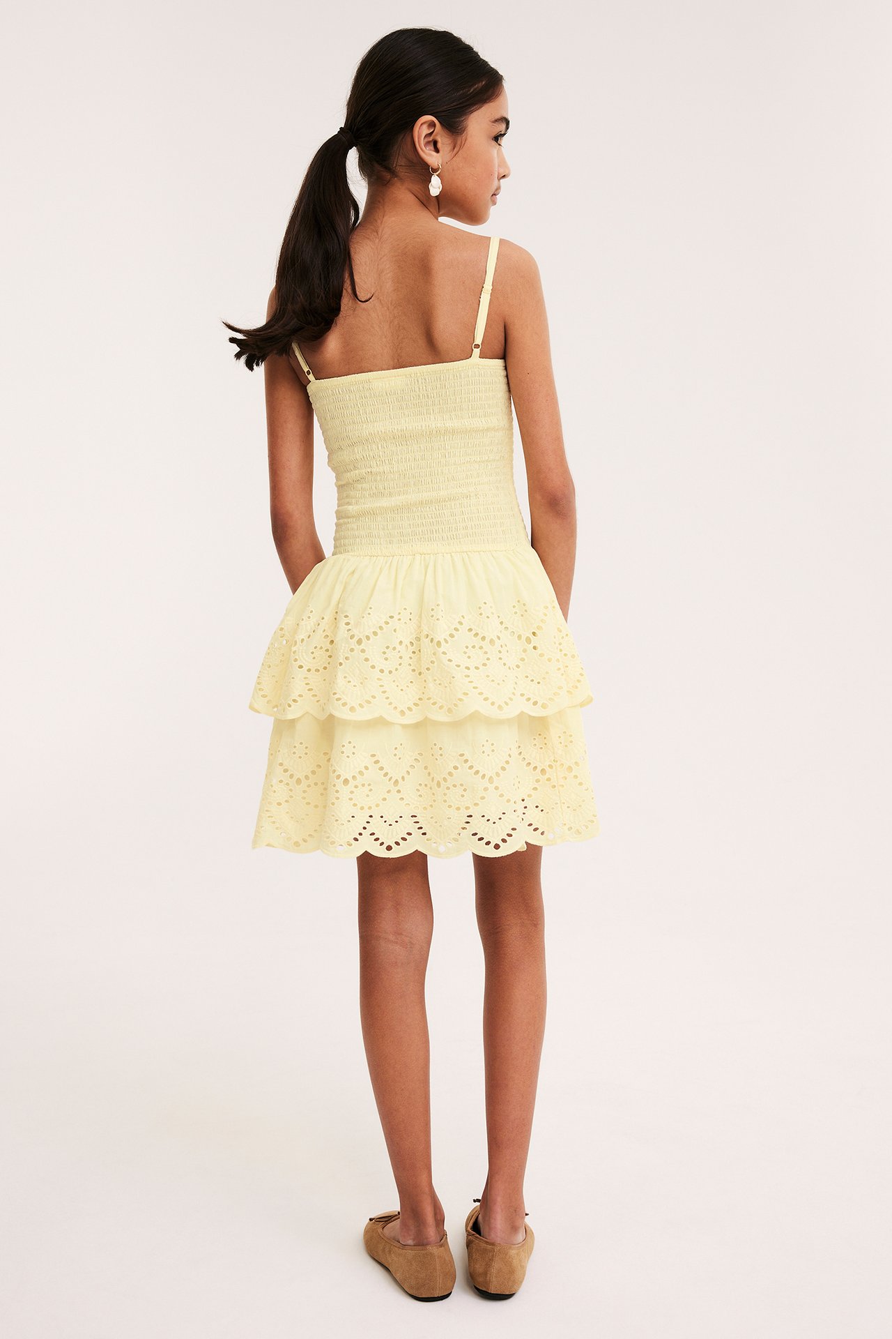 Bakifrån: barn i ljusgul smockad bomullsklänning, broderie anglaise-kjol i volanger, beiga platta skor, vita örhängen.