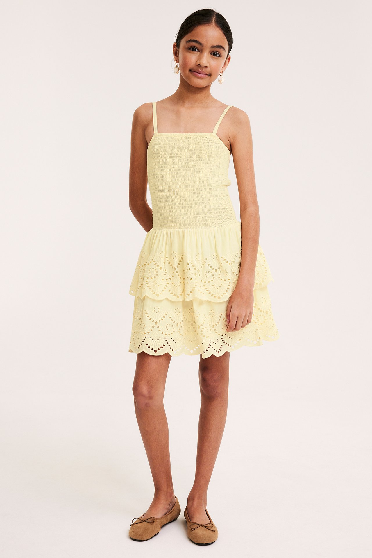 Framsida: Tjej i ljusgul smockad klänning med broderie anglaise-volanger och bruna ballerinaskor.