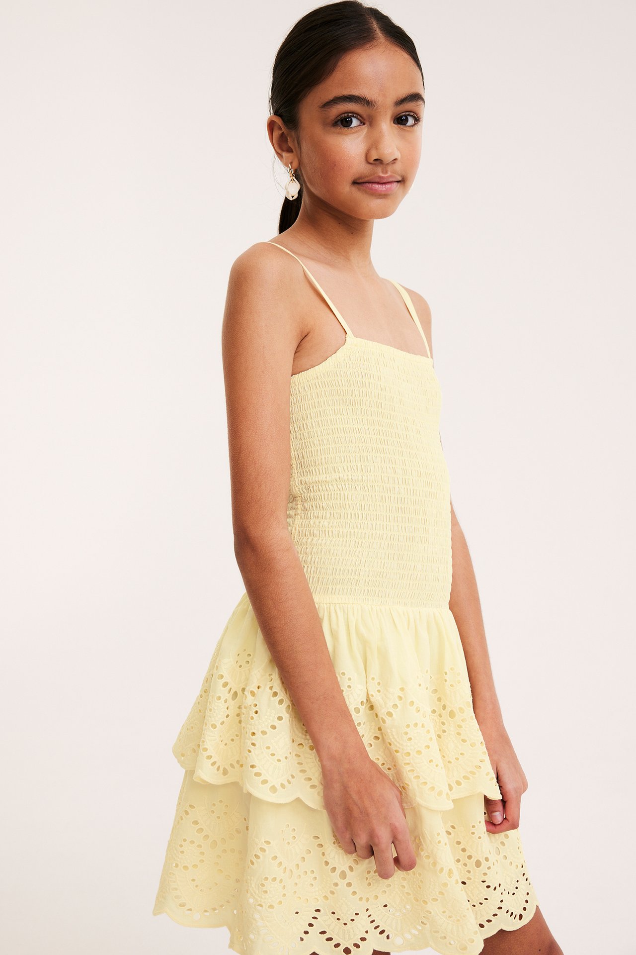 Front view: Kid i ljusgul smockklänning med broderie anglaise volanger, justerbara band. Bär vitt örhänge.