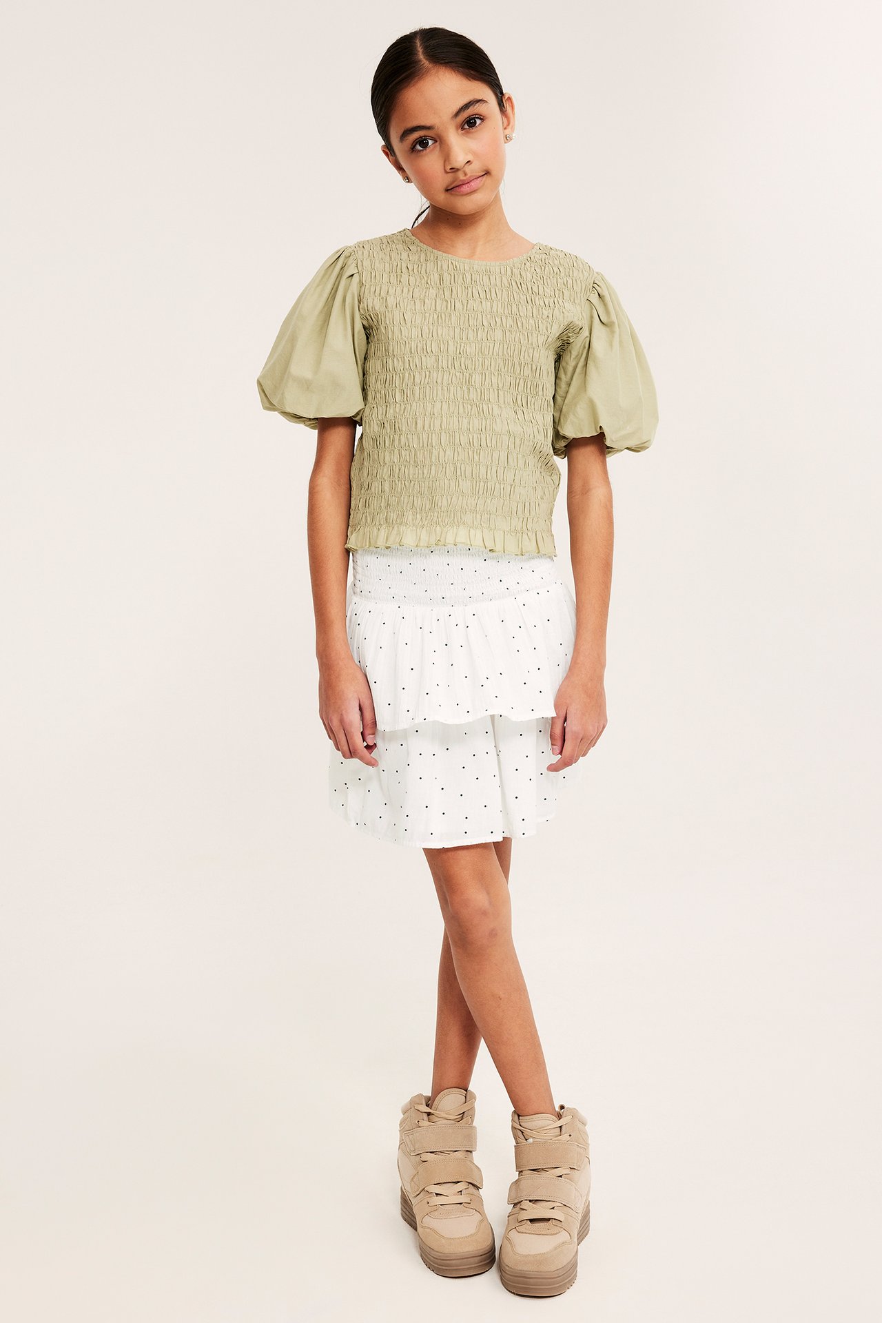 Framifrån: Barn bär vitprickig kort volangkjol i bomull, en olivfärgad smockad topp och beiga sneakers.