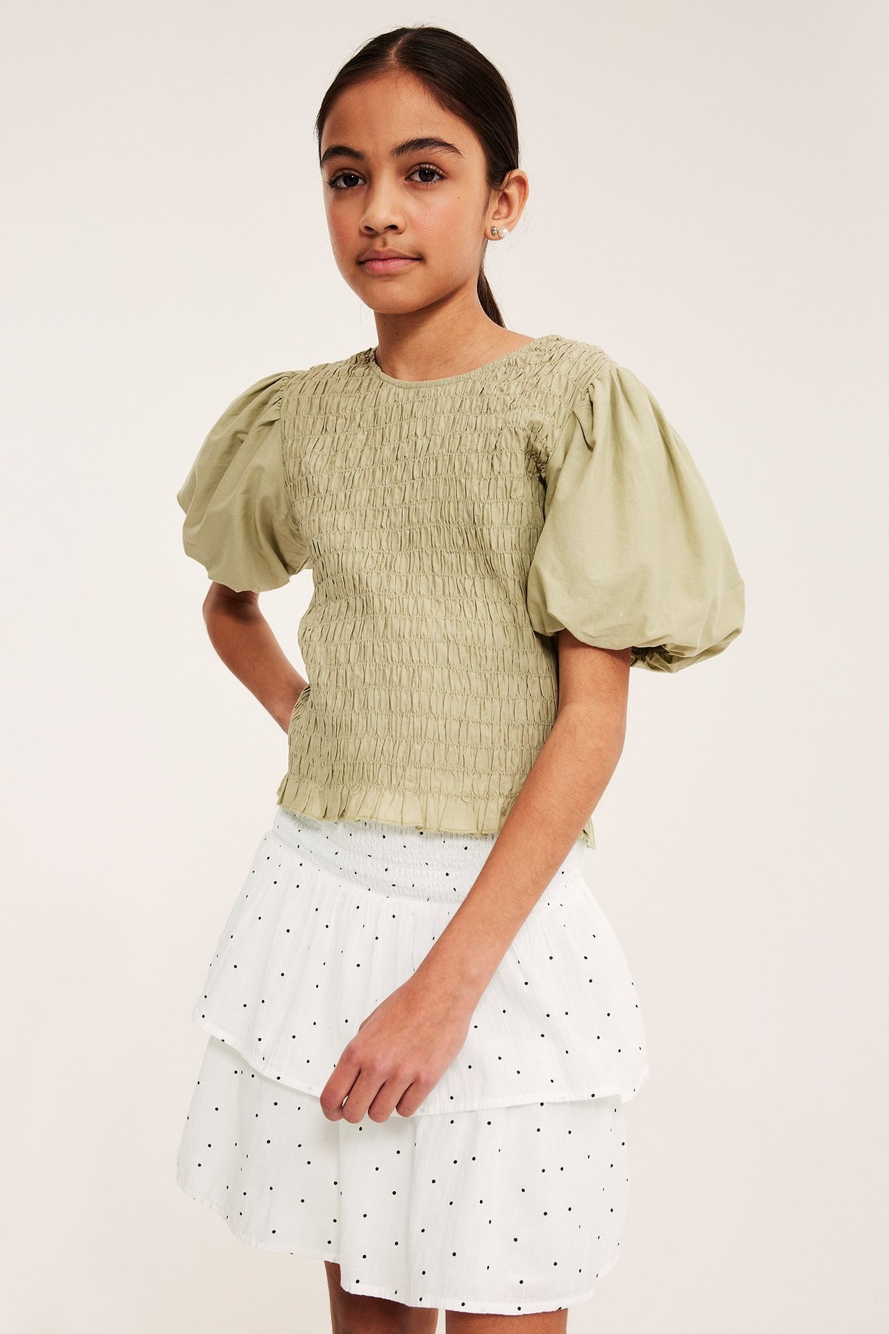Framifrån en flicka i vit prickig volangkjol med smock i midjan och olivgrön topp.