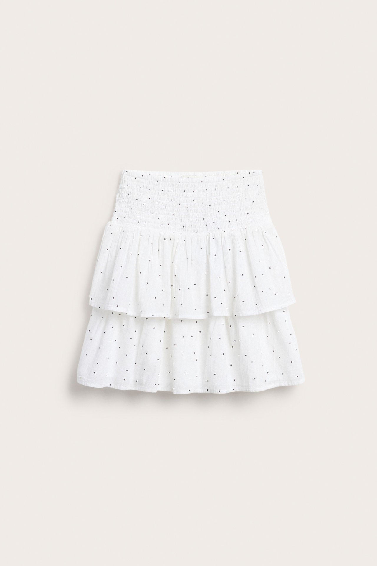 Edestäpäin kuvattu valkoinen puuvillainen lyhyt röyhelöhame, jossa smokkirypytys vyötäröllä ja mustia pilkkuja.