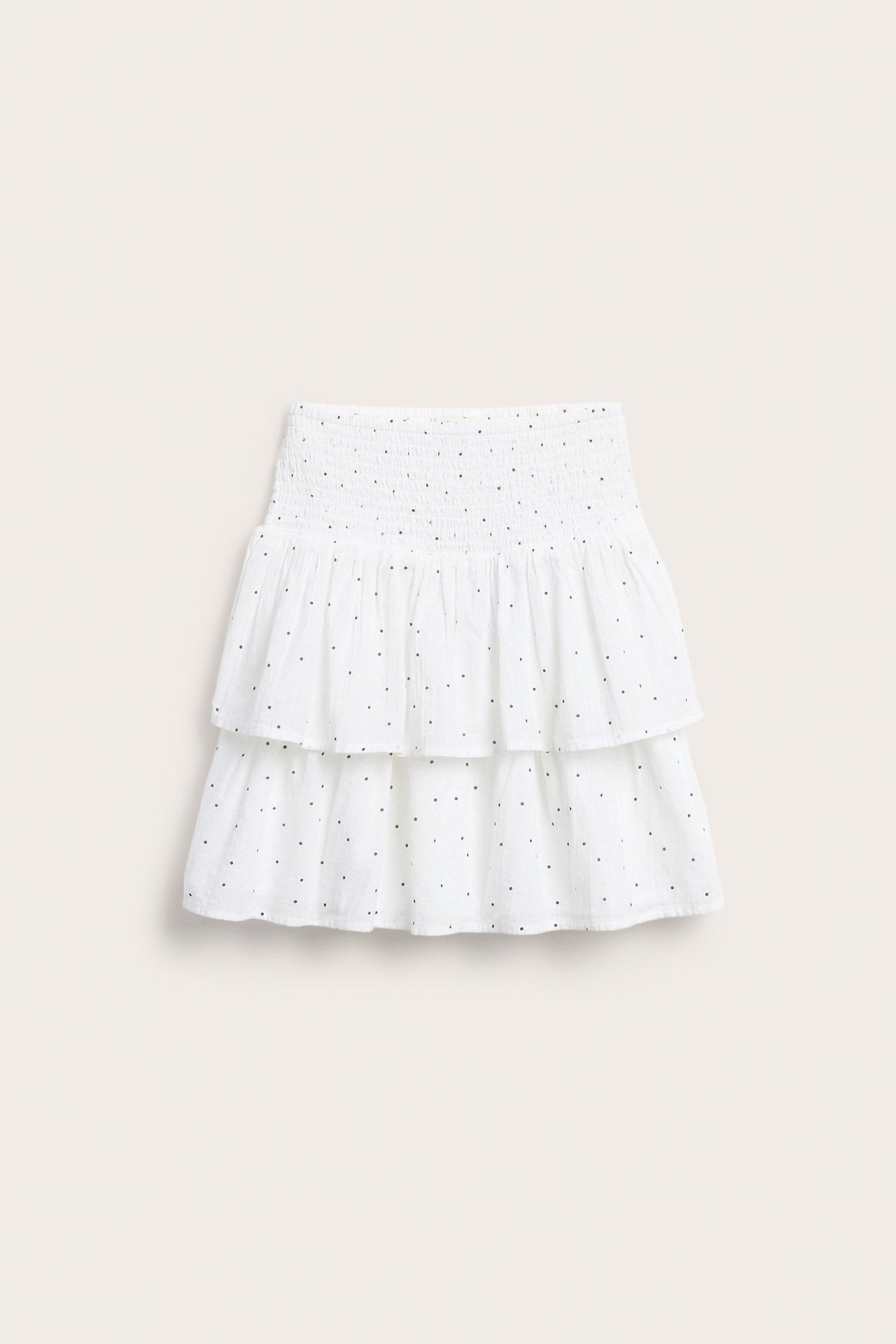 Frontvy av en vit kort volangkjol i bomull med smock i midjan och svarta prickar.