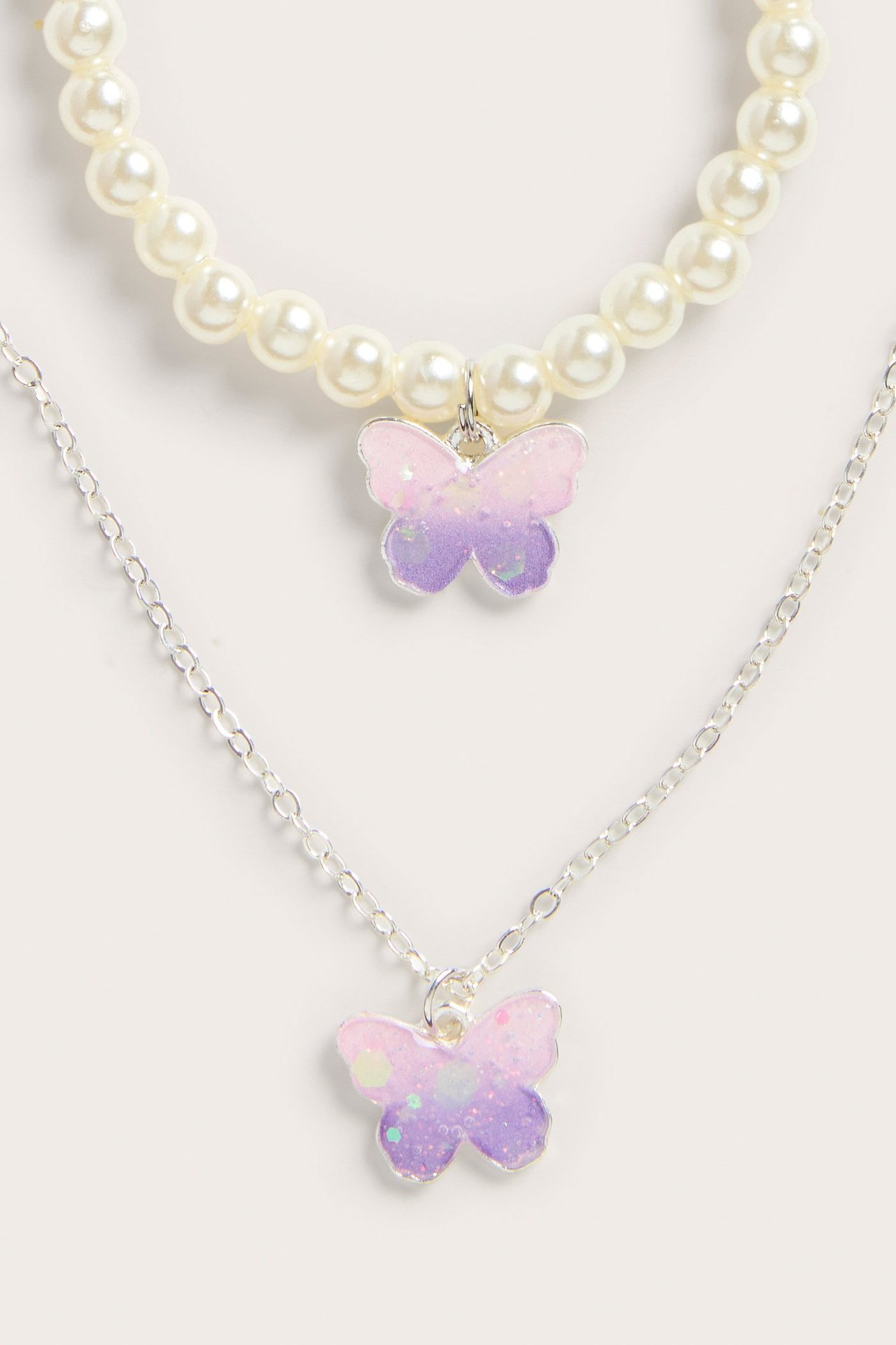 Nærbilde: et hvitt perlekjede og en sølvfarget kjede, begge med et rosa-lilla glitter-sommerfuglanheng.