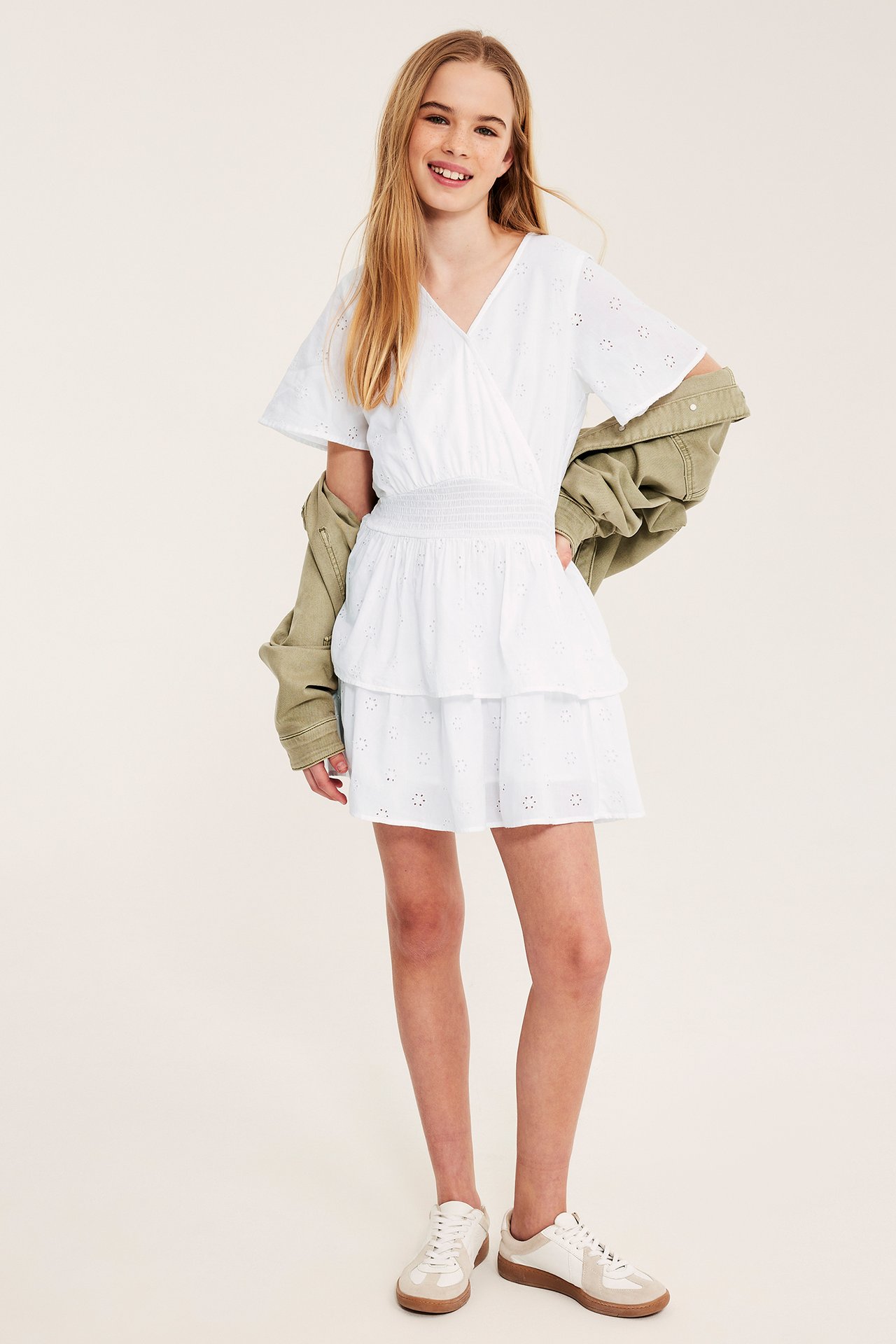 Edestäpäin: Tyttö valkoisessa pitsikuvioidussa puuvillamekossa, khakinvärinen takki ja valkoiset tennarit.