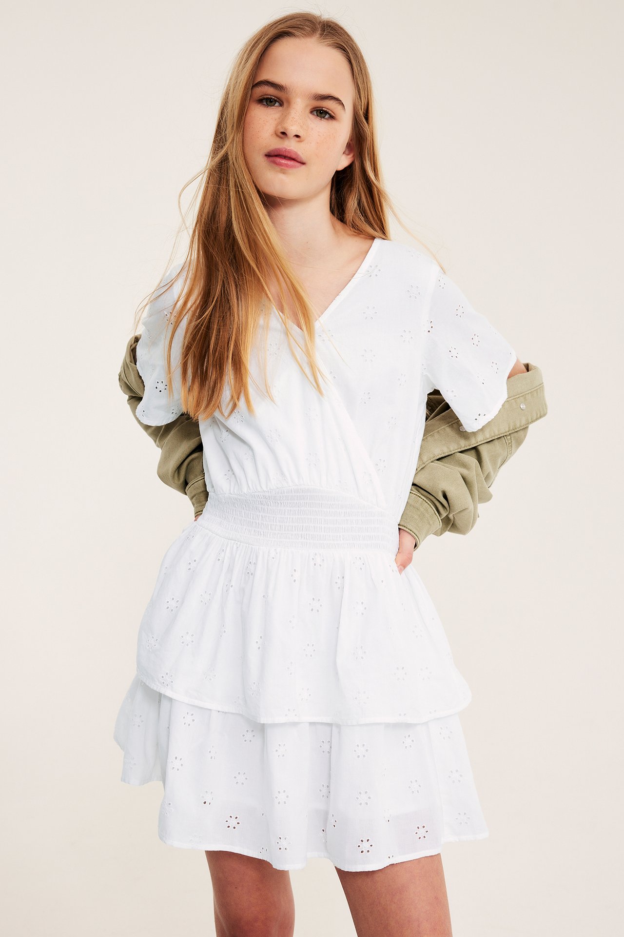 Dziewczynka w białej bawełnianej sukience z haftem angielskim, marszczoną talią i falbankami, z kurtką khaki.