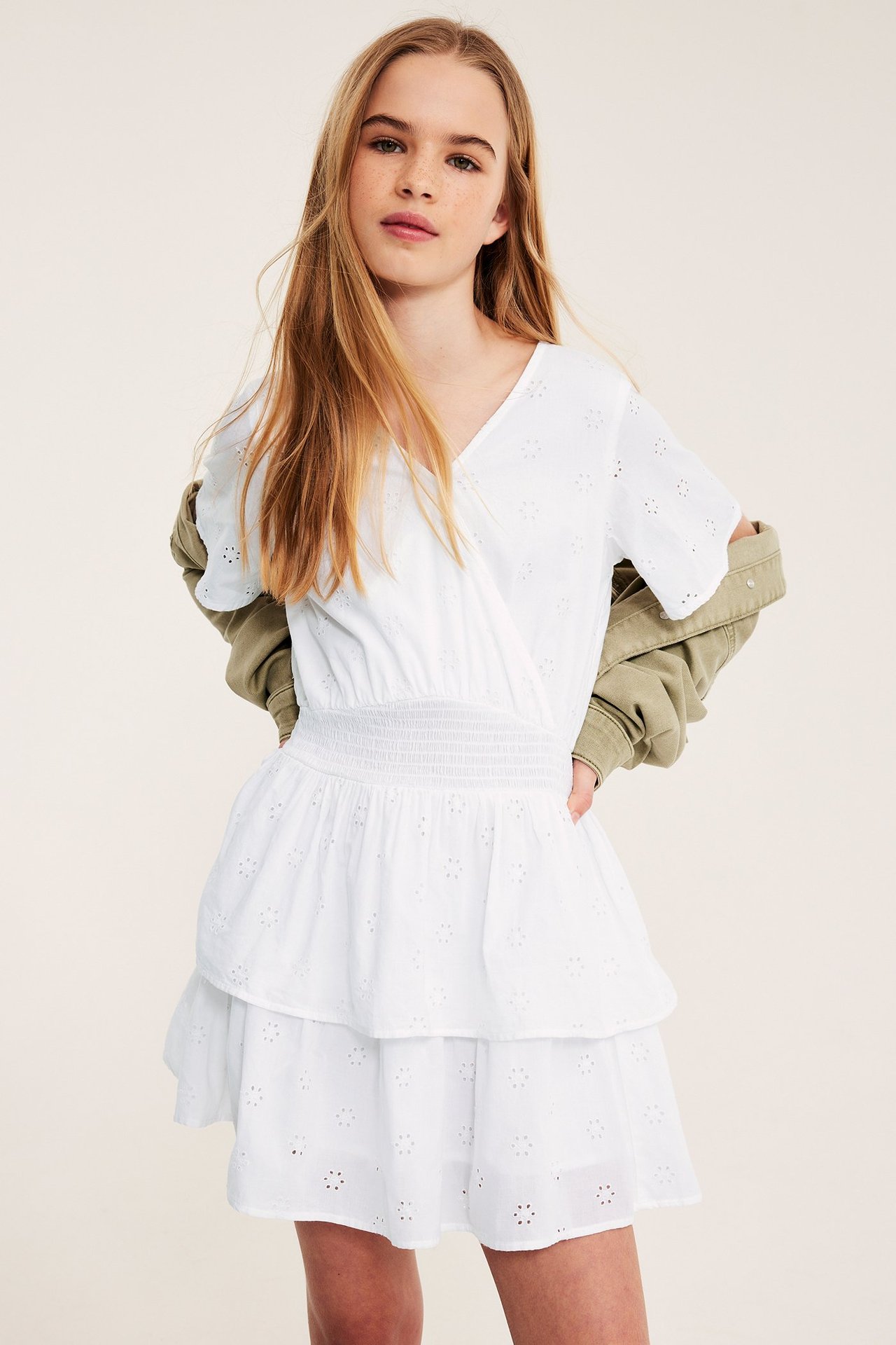 Barn i hvit bomullskjole med broderie anglaise, smock i midjen og volanger, med en khaki jakke.