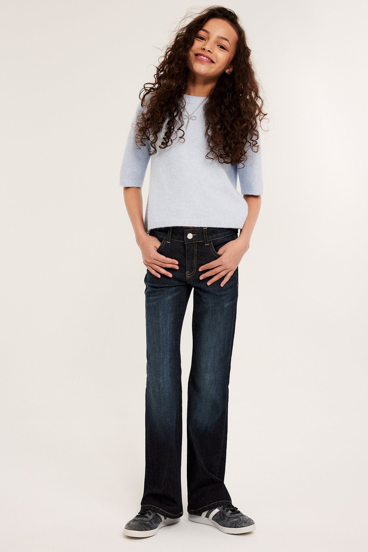 Frontvy av ett barn i mörkblå bootcut jeans med låg midja, ljusblå kortärmad tröja och mörka sneakers.