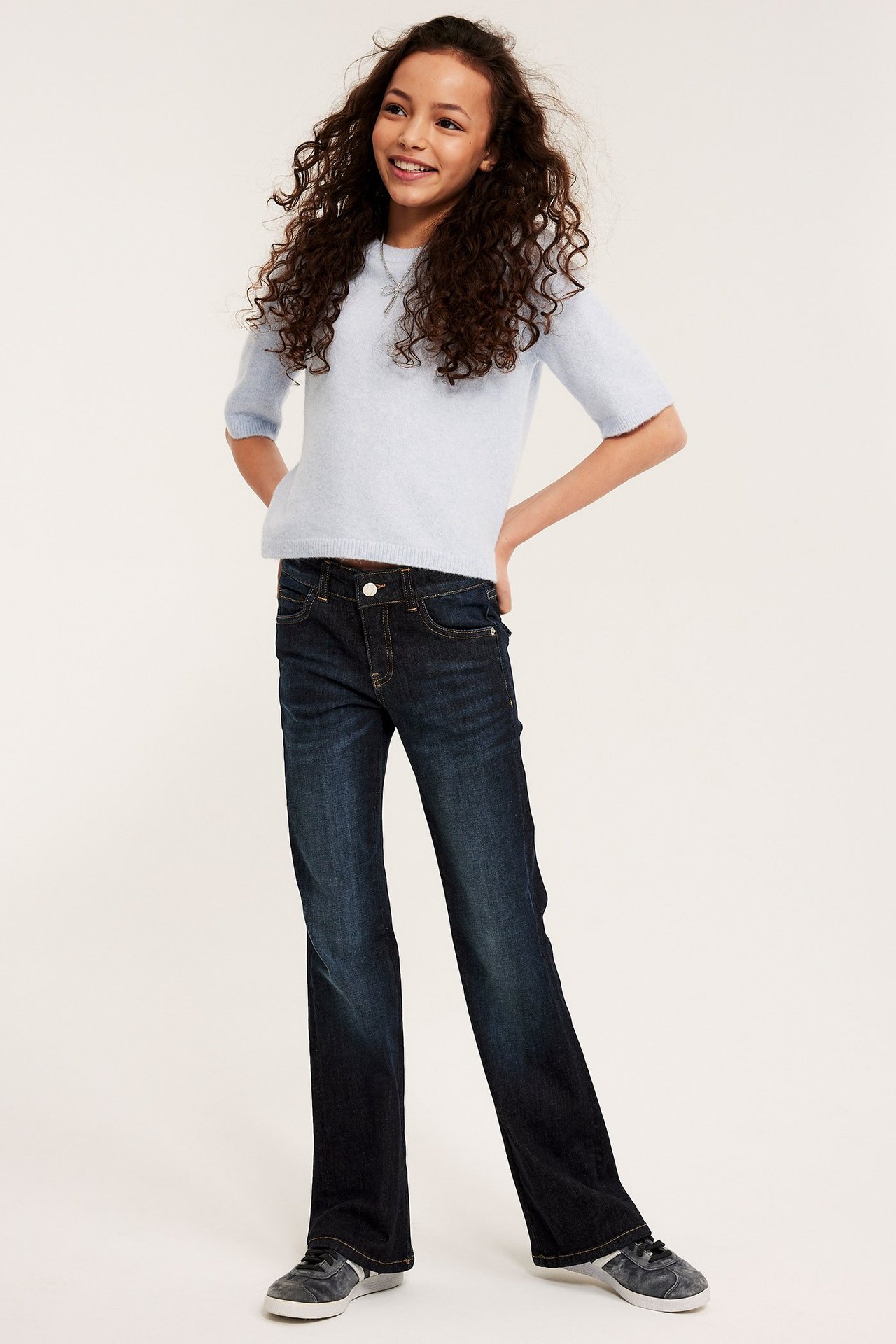 Widok z przodu: Uśmiechnięte dziecko w ciemnoniebieskich jeansach bootcut z niskim stanem, jasnoniebieskim swetrze i ciemnoszarych sneakersach.