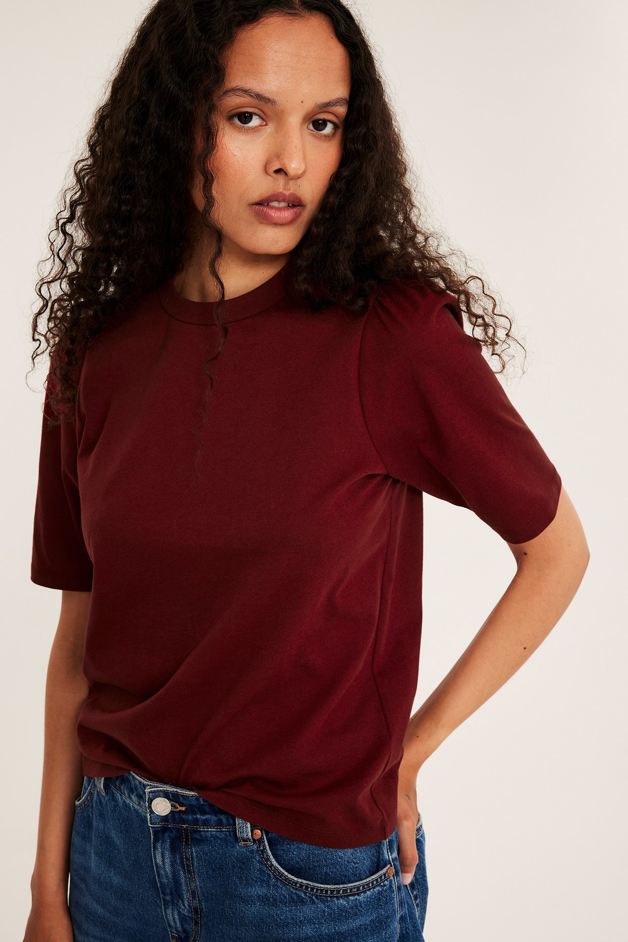 Widok z przodu: kobieta w bordowym, jednokolorowym topie bawełnianym z bufiastymi rękawami i niebieskich dżinsach.