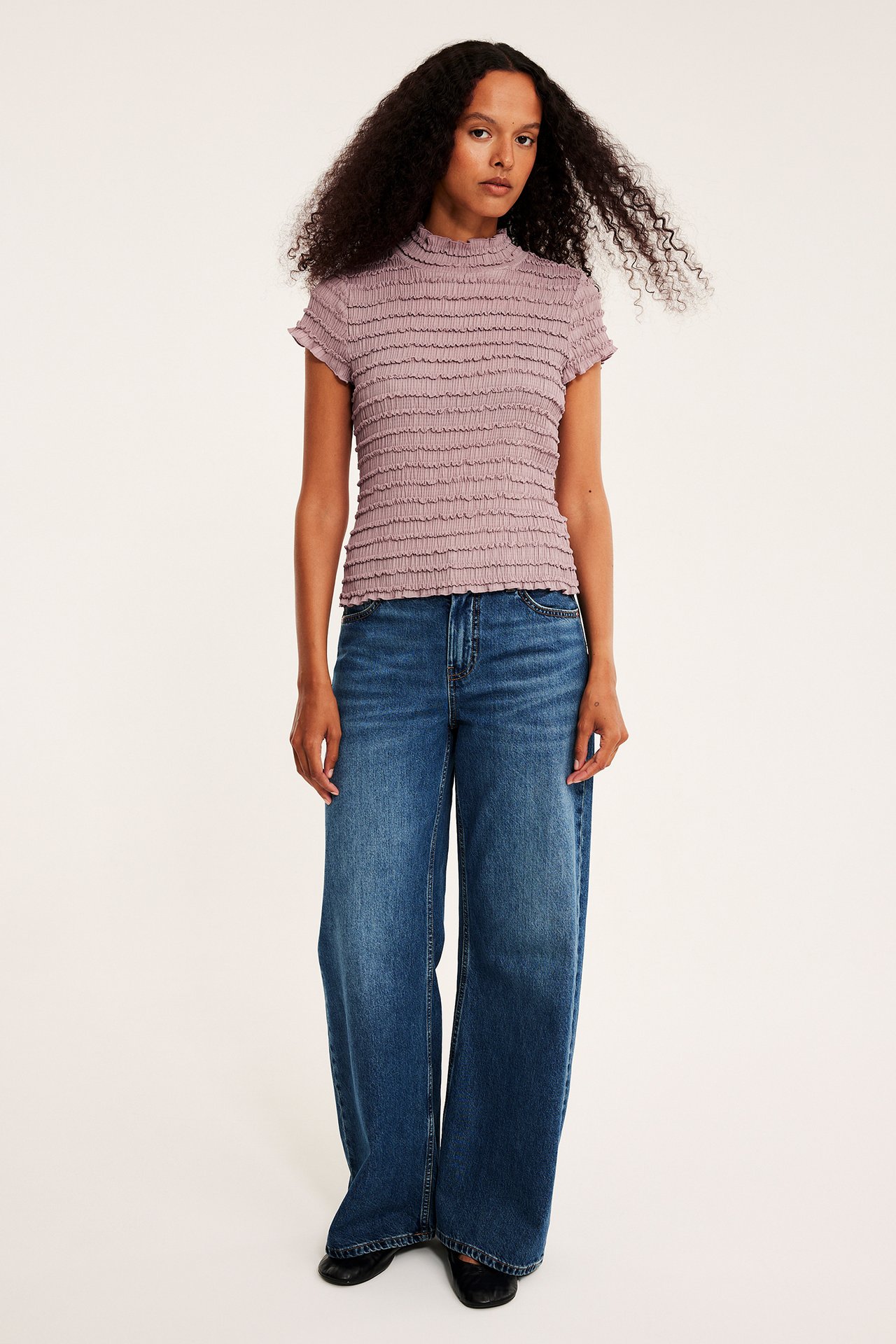 Kvinnelig modell i lilla rynket høyhalset topp, blå vide jeans og svarte flate sko.