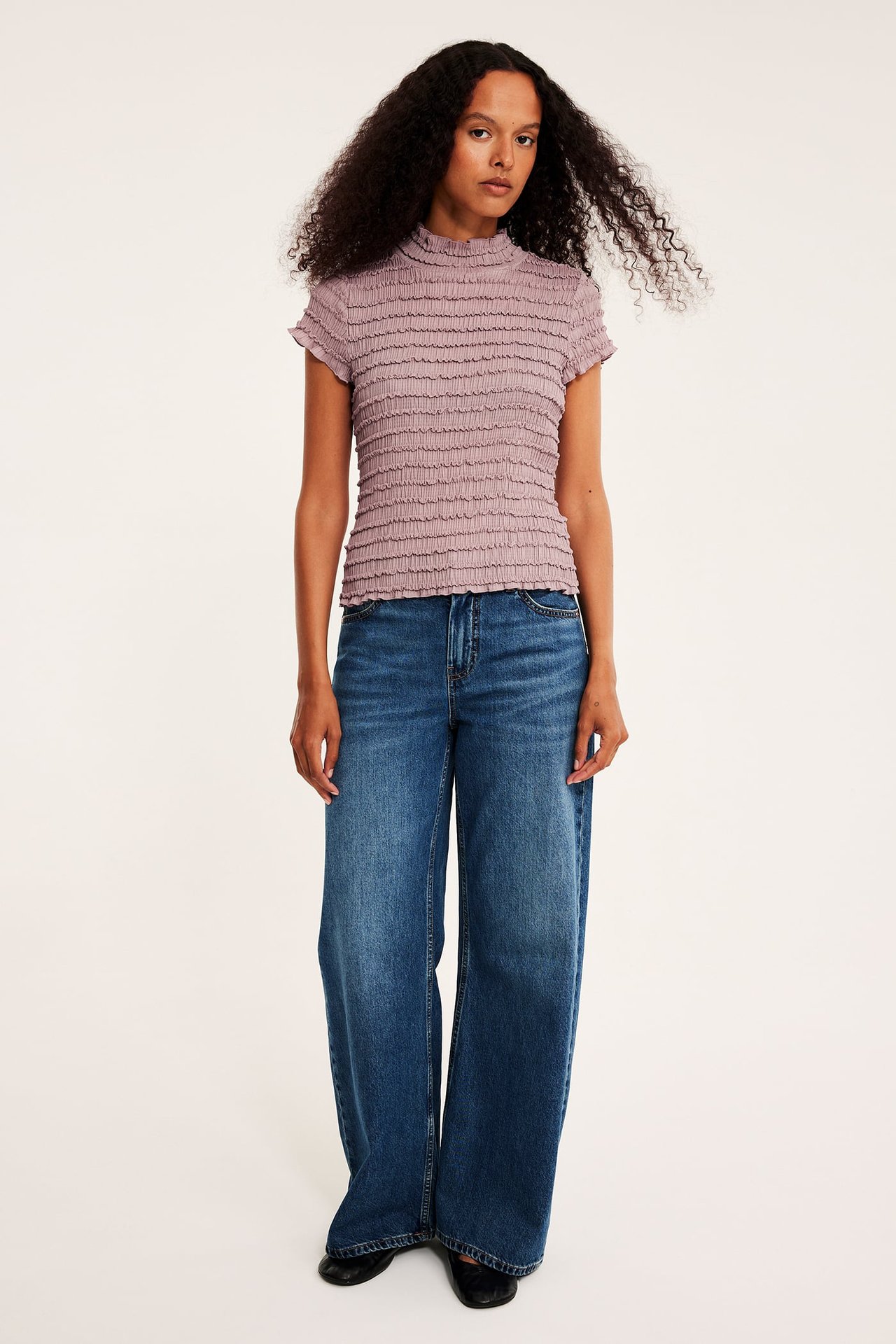 Kvinnelig modell i lilla rynket høyhalset topp, blå vide jeans og svarte flate sko.