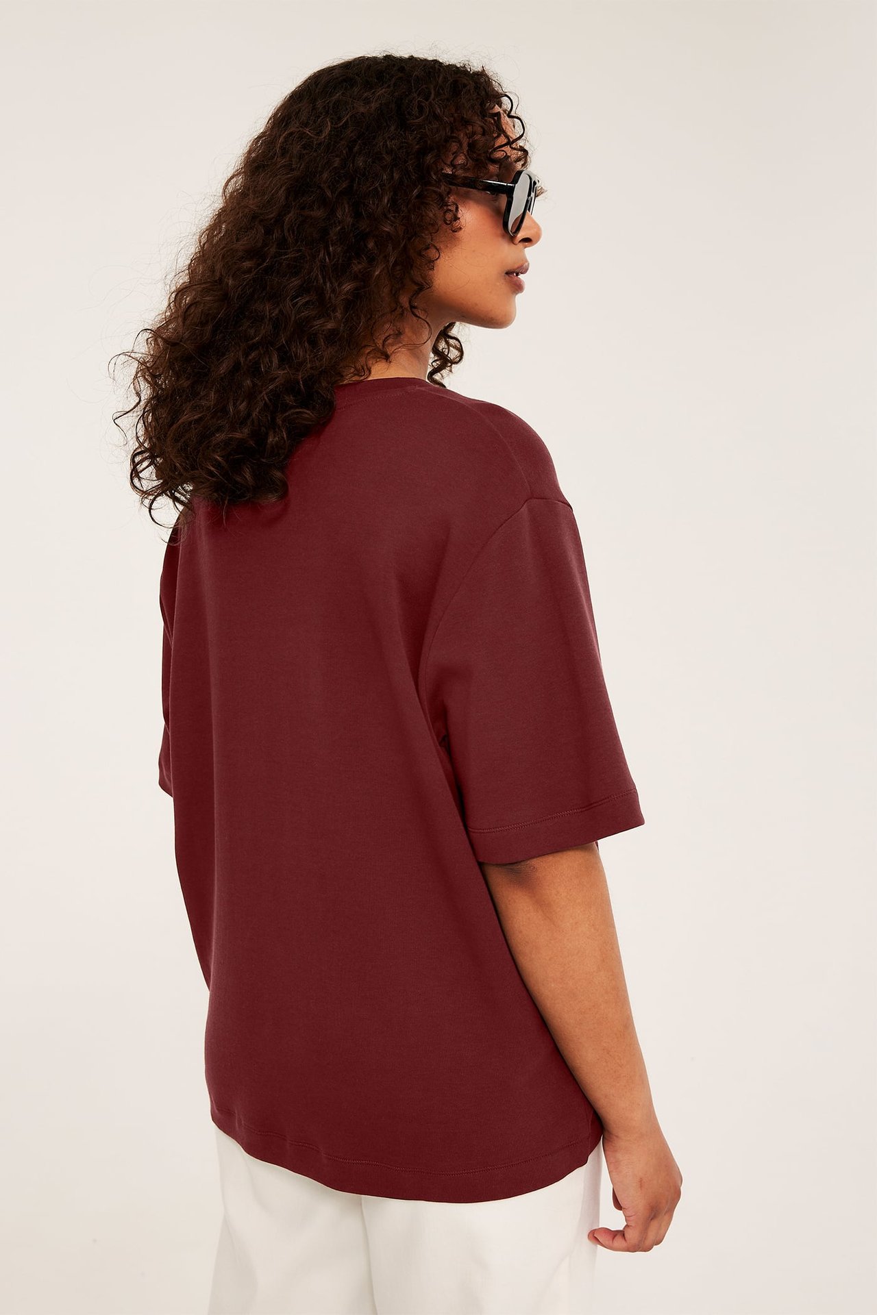 Sidovy av kvinnlig modell i en vinröd oversized t-shirt, vita byxor och svarta solglasögon.