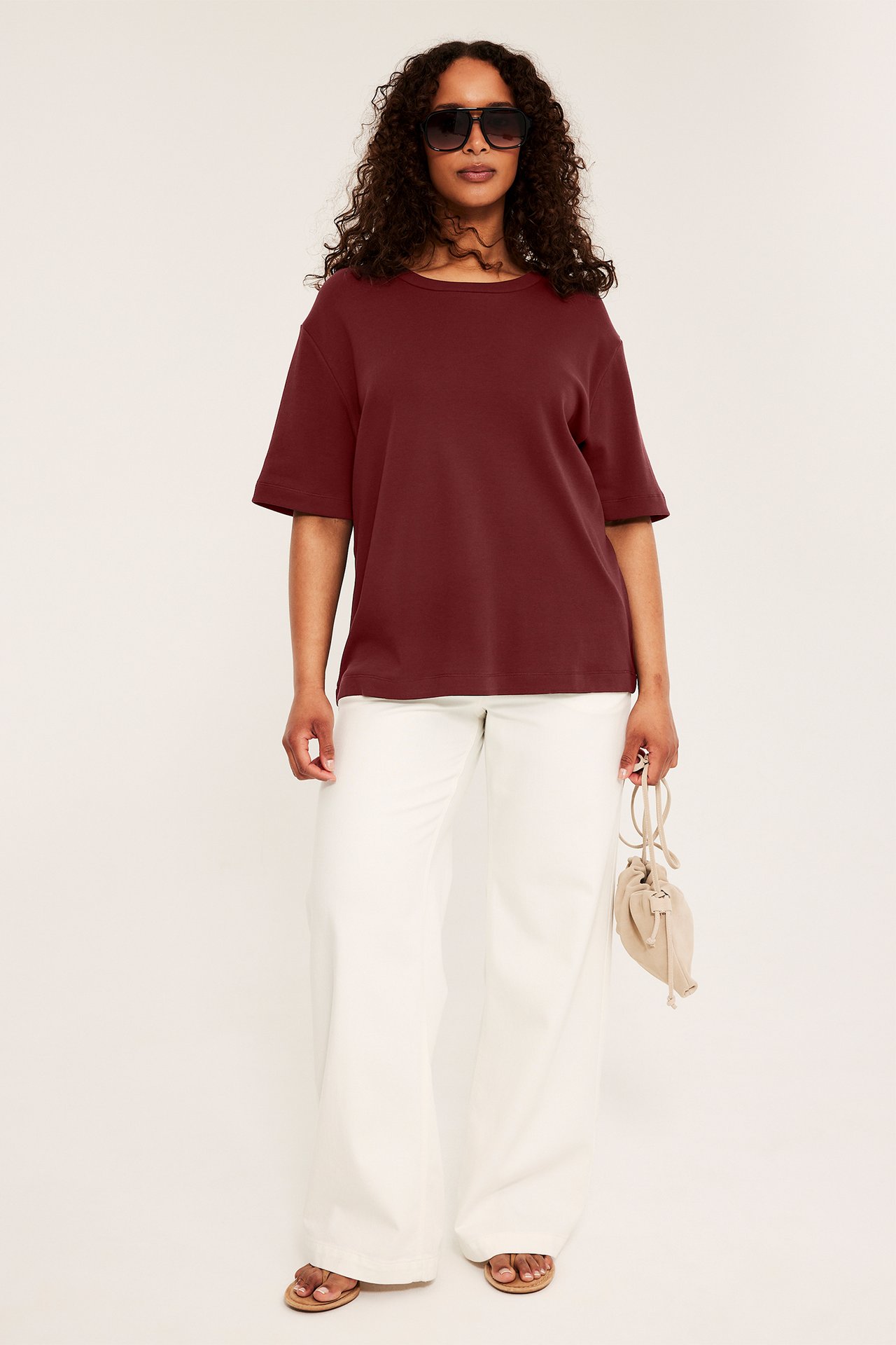 Framifrån kvinna i vinröd t-shirt, vita vida byxor, bruna sandaler, håller beige väska.
