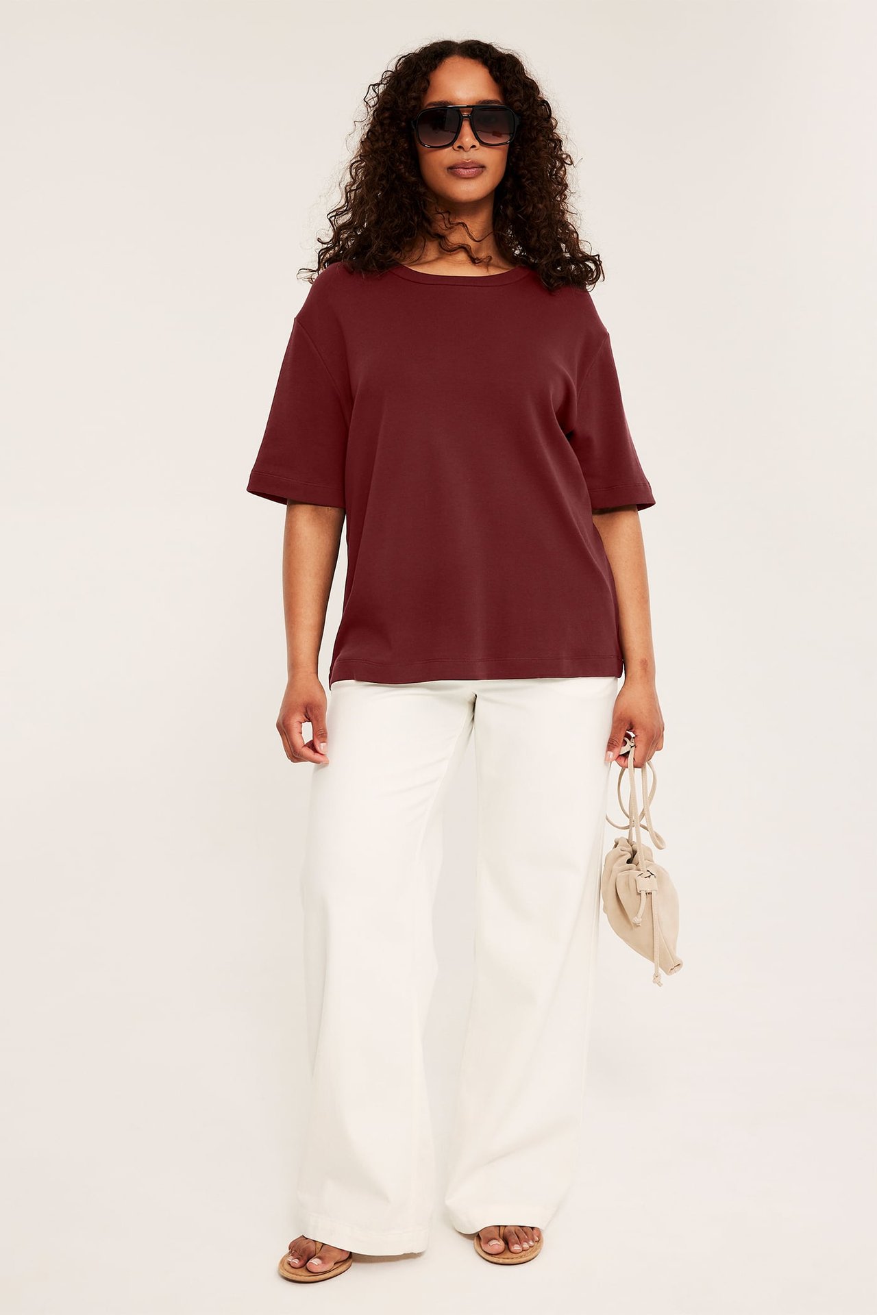 Framifrån kvinna i vinröd t-shirt, vita vida byxor, bruna sandaler, håller beige väska.