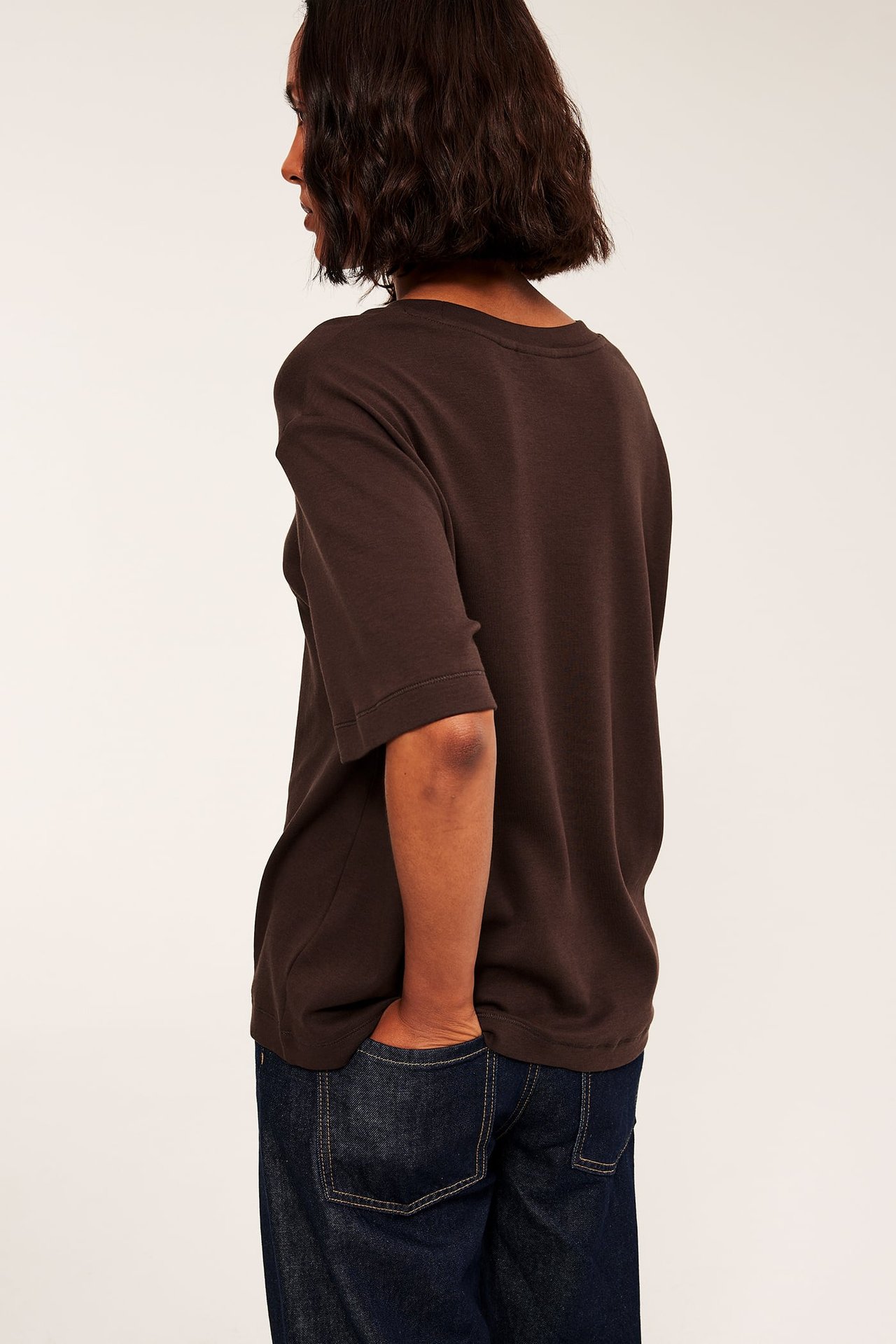 Bakre sidovy av en vuxen kvinna som bär en brun kortärmad t-shirt och mörkblå jeans.