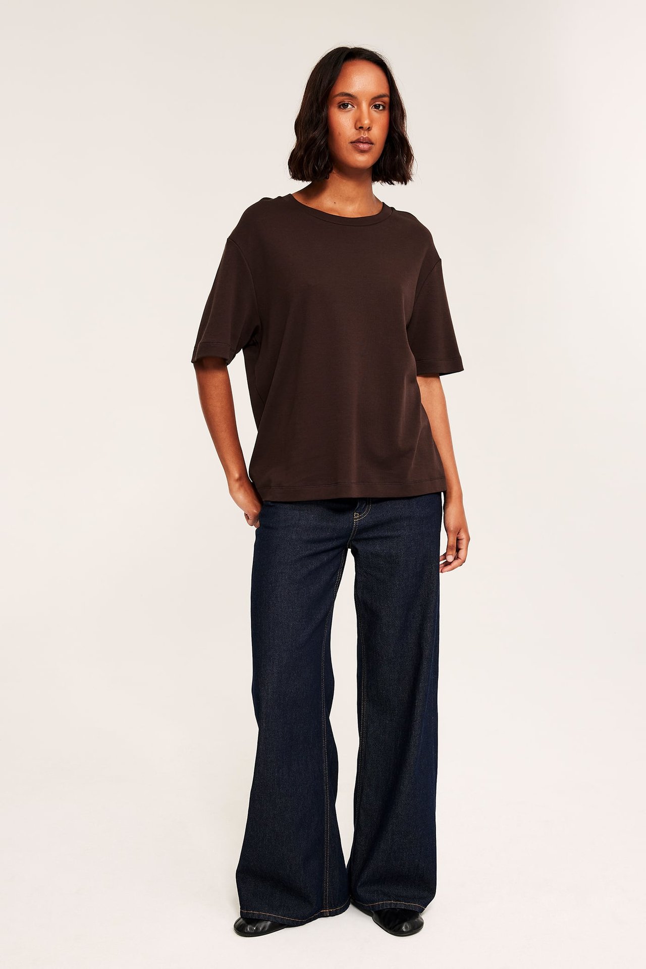 Front vy av en kvinna i en brun oversized t-shirt, mörkblå vida jeans och svarta platta skor.