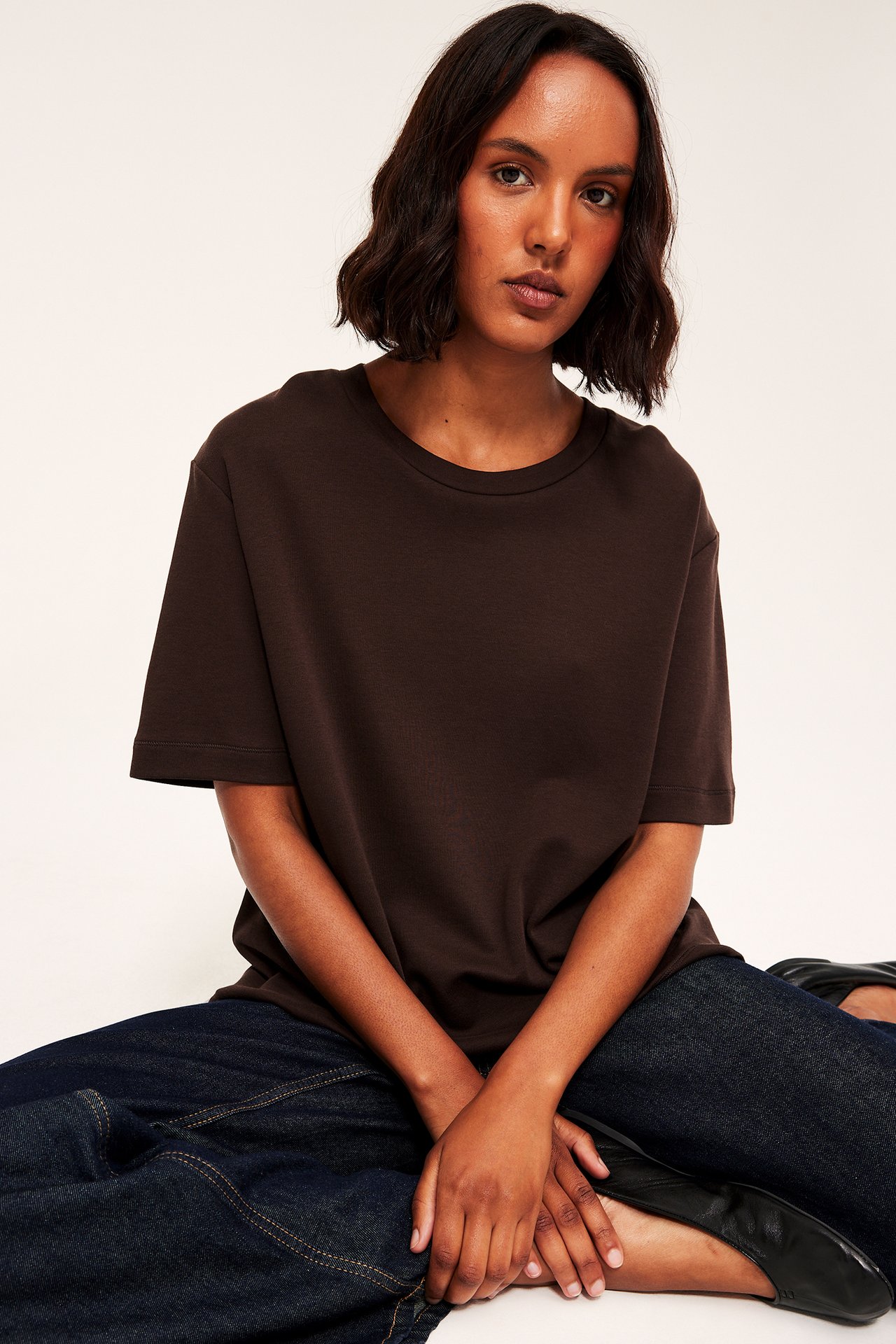 Framifrån, kvinna i brun vid t-shirt, mörkblå jeans och svarta ballerinaskor.