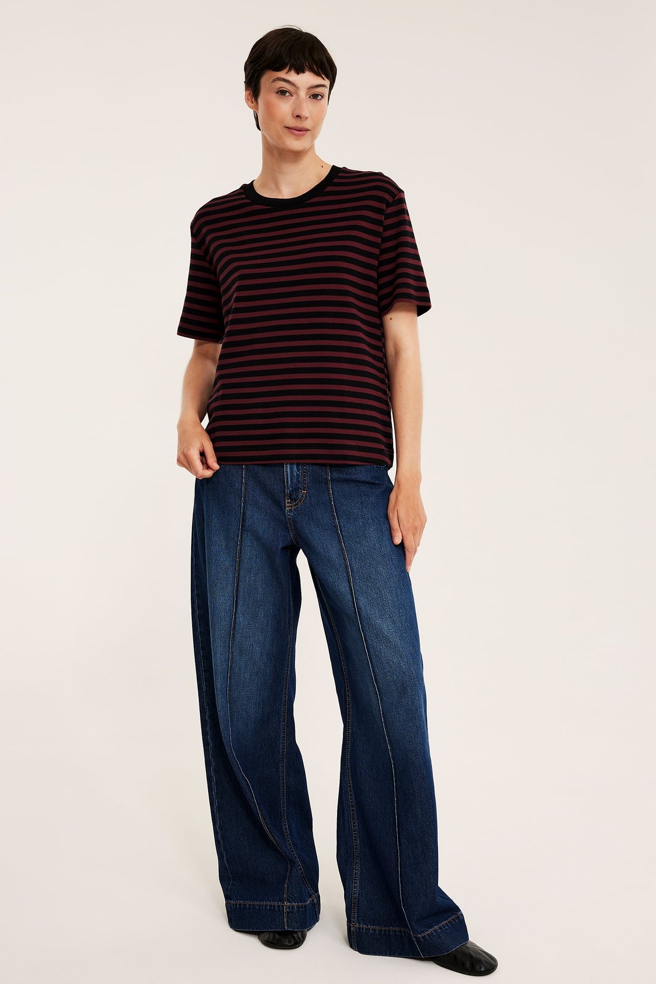 Front view av en kvinnlig modell i svart och vinrödrandig t-shirt och vida blå jeans.