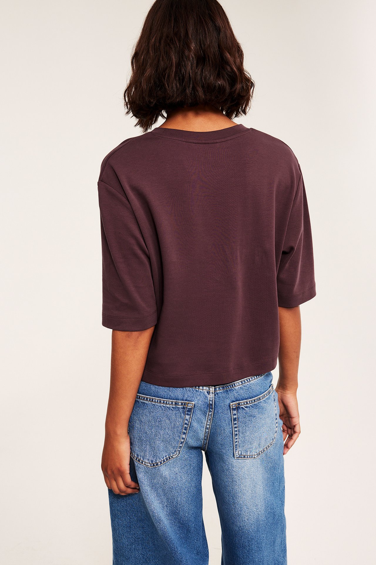 Bakifrån: kvinna i mörk vinröd kortärmad croppad t-shirt och blå denimjeans.