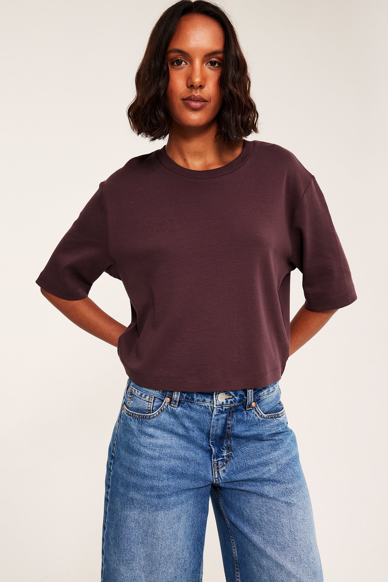 Frontvy av en kvinna i mörkbrun kortärmad topp och blå vida jeans.