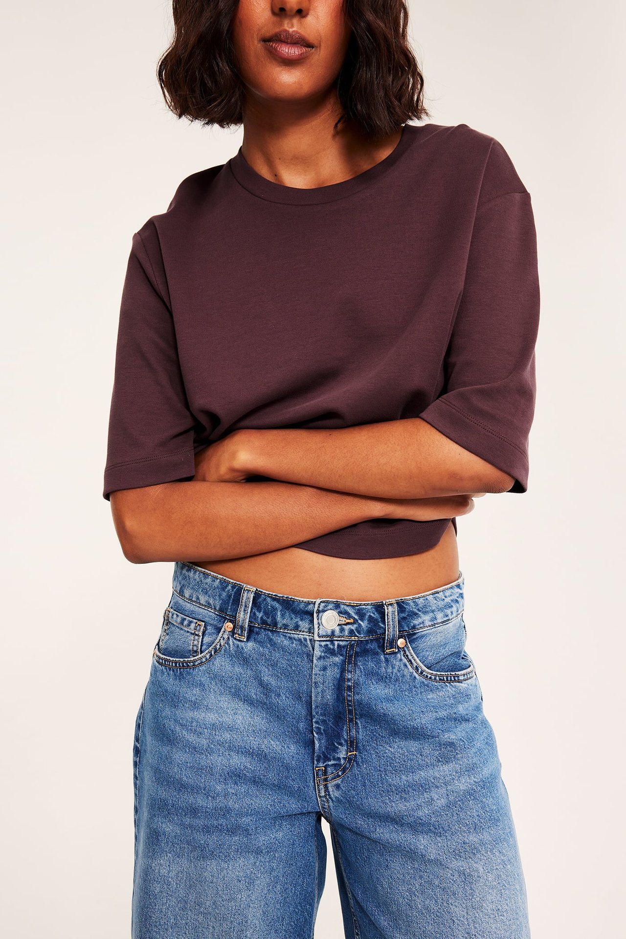 Frontvy av kvinna i mörk plommonfärgad croppad t-shirt med trekvartsärm och blå jeans.