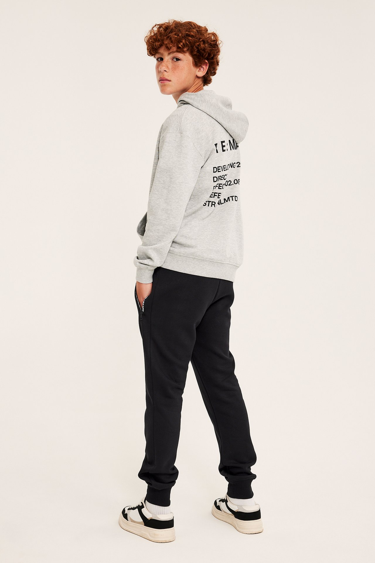 Ryggvy av ett barn i grå hoodie med svart text, svarta mjukisbyxor och svartvita sneakers.