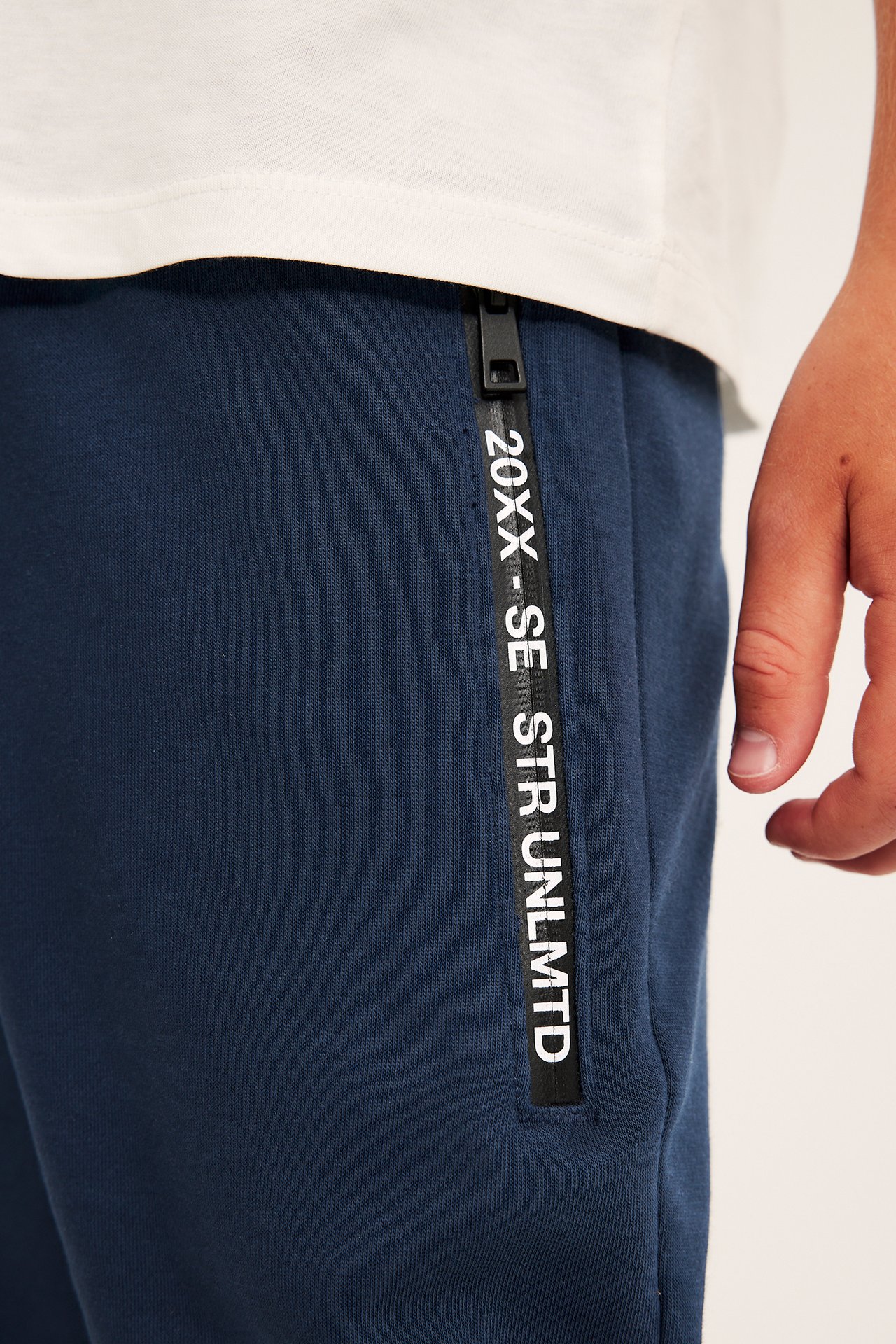 Närbild av mörkblå mjukisbyxor med svart textsidrand och dragkedja, buren med vit t-shirt.