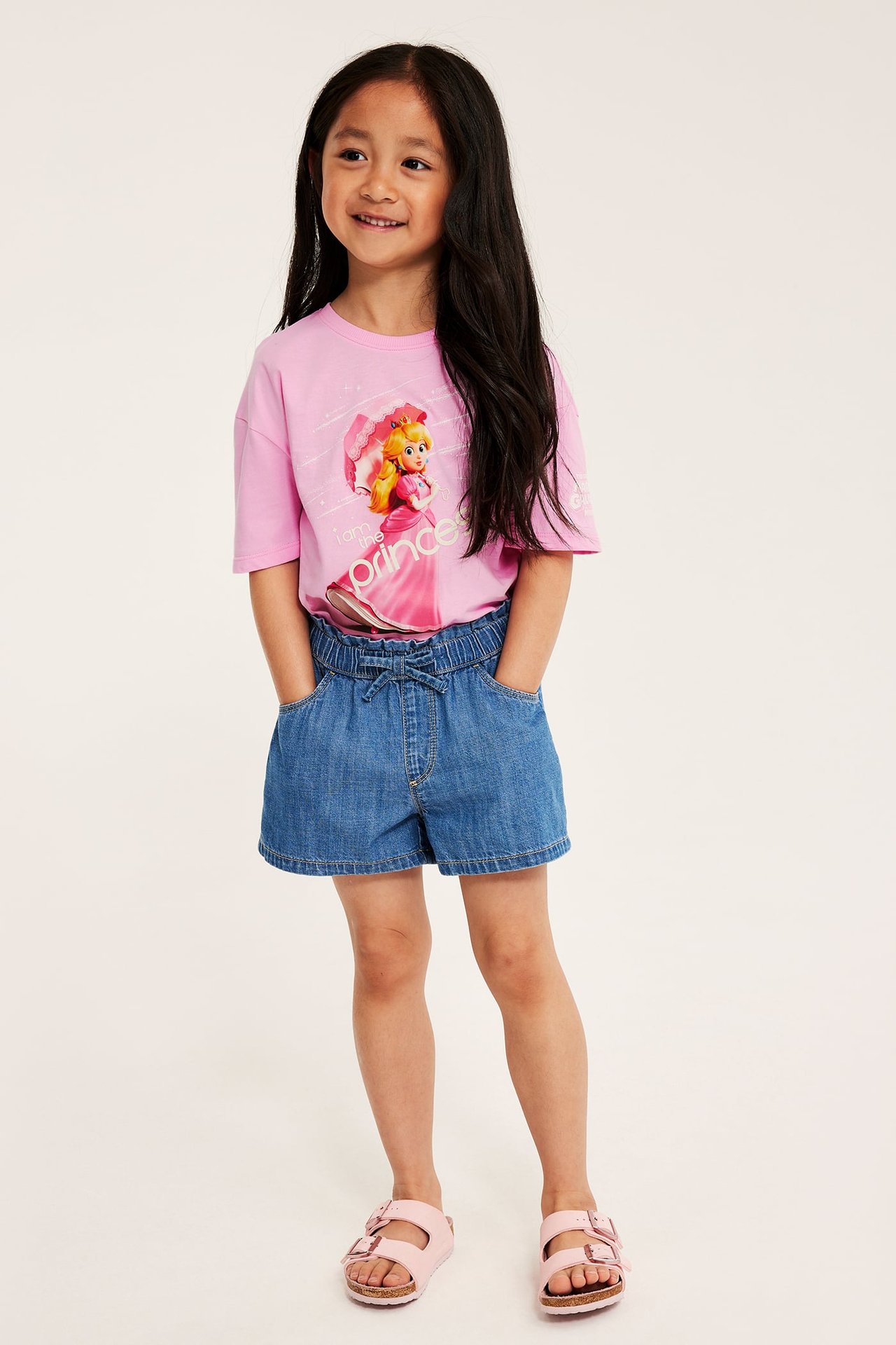 Leende barn bär ljusrosa bomullst-shirt med Prinsessan Peach-tryck, blåa jeansshorts och rosa sandaler.