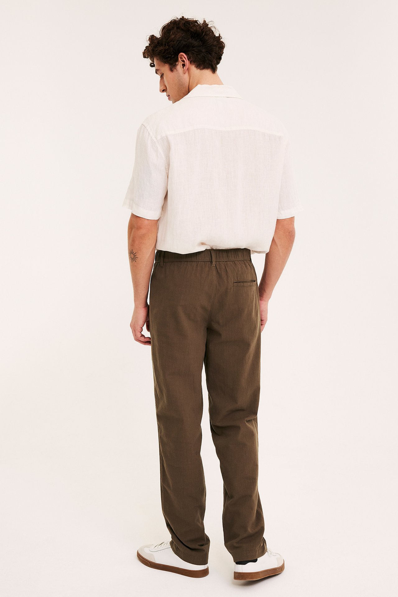 Widok z tyłu: mężczyzna w brązowych bawełnianych spodniach o wyglądzie lnu z gumką, białej koszuli i białych sneakersach.