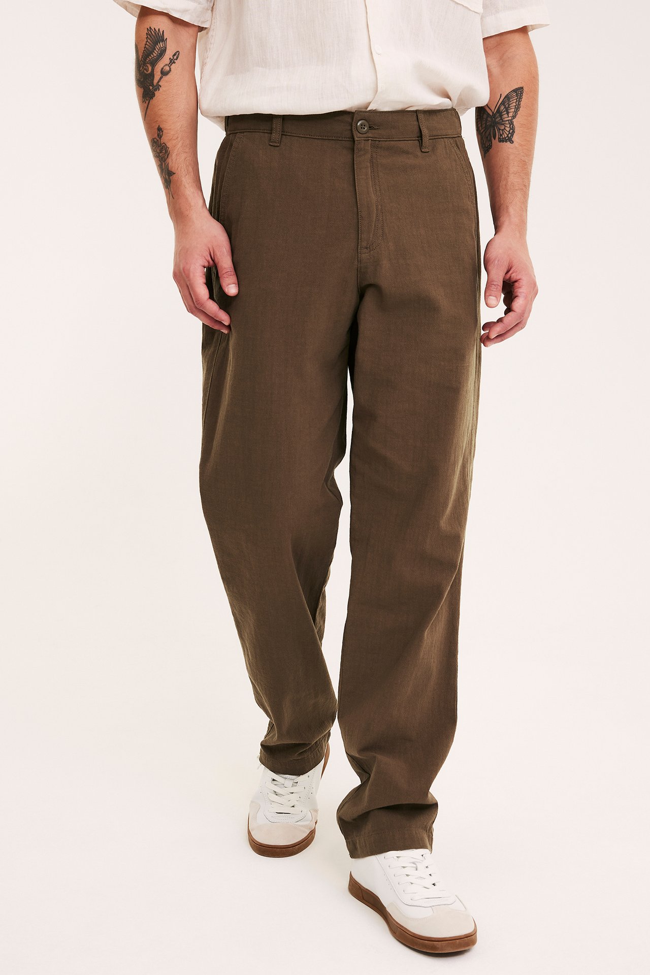 Mann forfra i brun bomullsbukse med linnenlook, en lys kortermet skjorte og hvite joggesko.