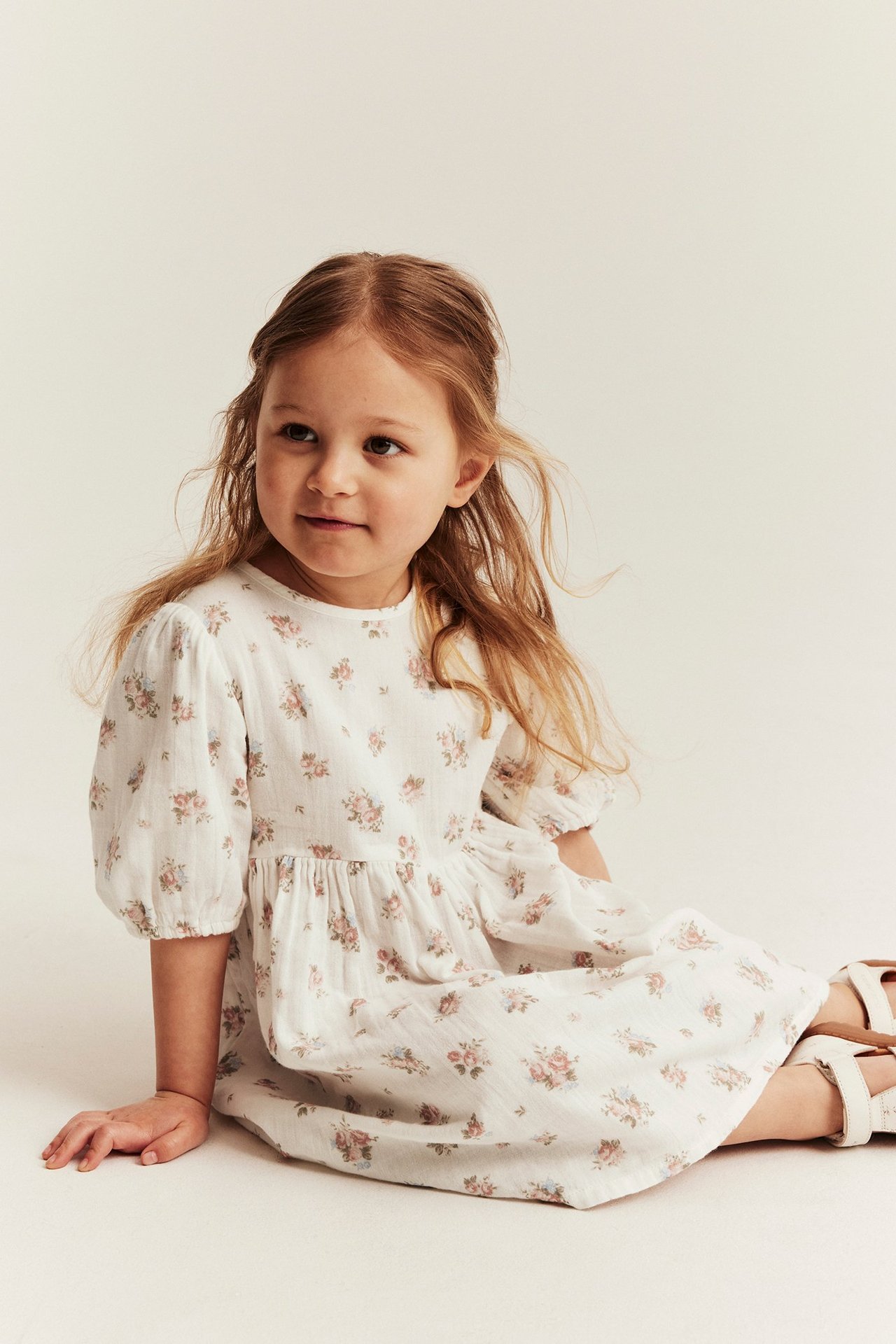 Widok z przodu dziecka w kremowej, bawełnianej sukience w kwiaty z bufiastymi rękawami i białych sandałach.