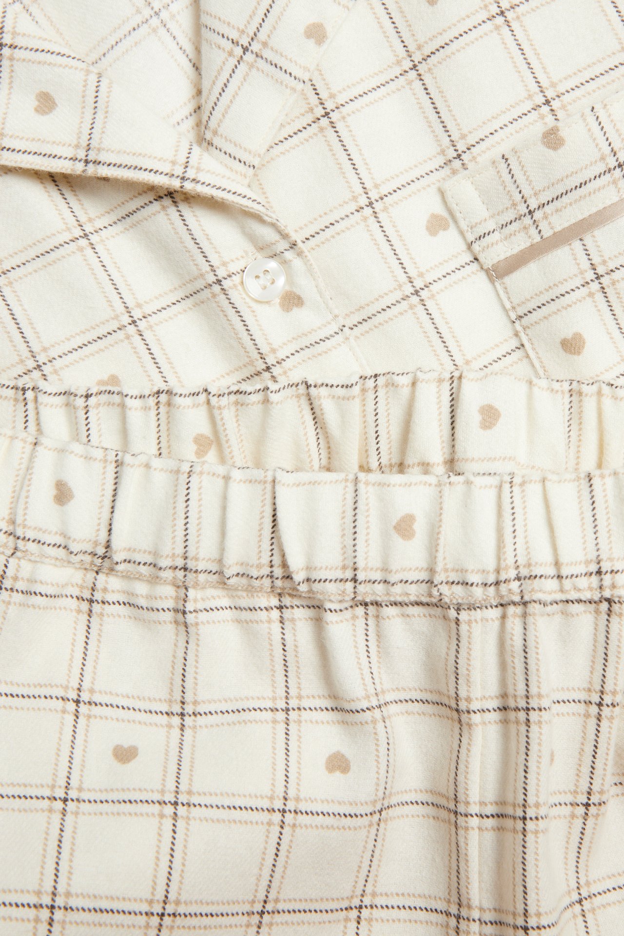 Lähikuva kermanvärisestä ruudullisesta pyjamasta ruskeilla sydämillä, napitettava yläosa ja kuminauhahousut.