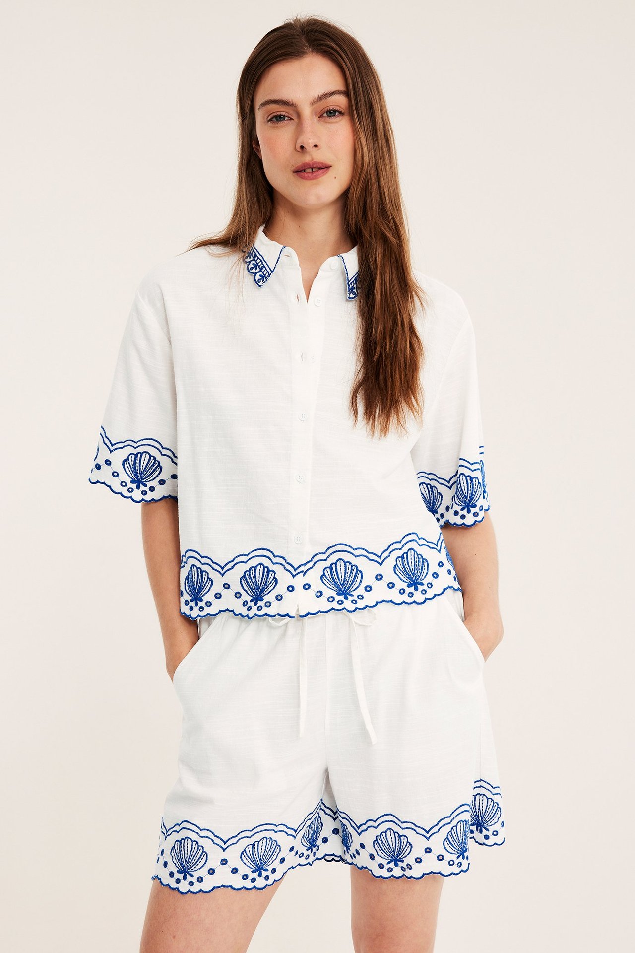 Framifrån: Kvinna i offwhite bomullsshorts med blått snäckbroderi och matchande kortärmad skjorta.