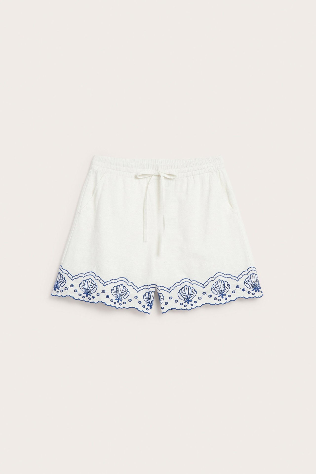 Framifrån vita bomullsshorts med dragsko, sidfickor och blå snäckmönstrad broderie anglaise fåll.