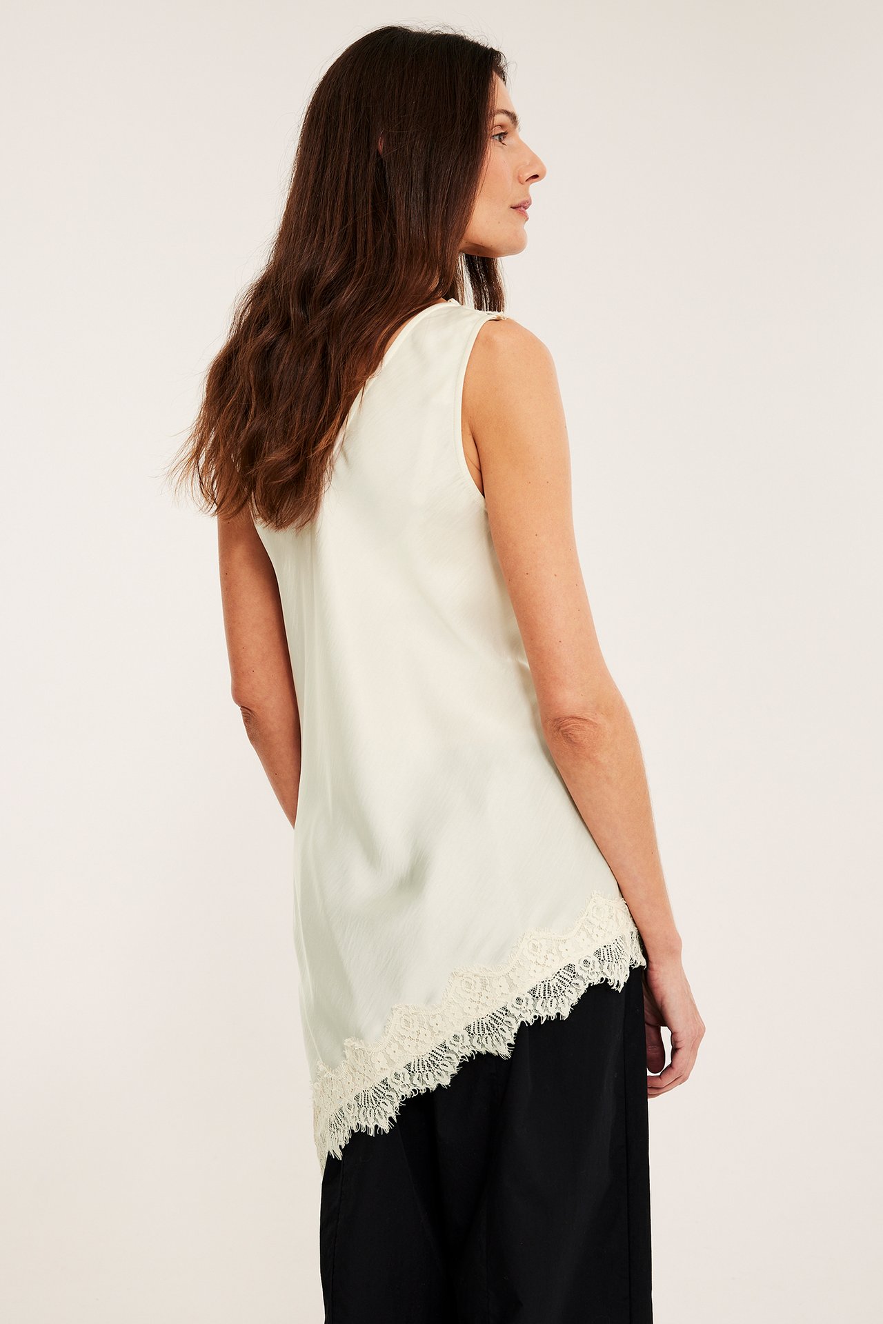 Widok z boku kobiety w écru satynowym topie z asymetrycznym koronkowym dołem, czarne spodnie.