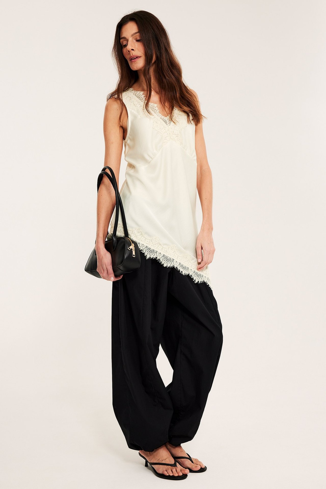 Kvinna i offwhite linne i satin med spets, svarta vida byxor och svarta sandaler, håller en svart handväska.