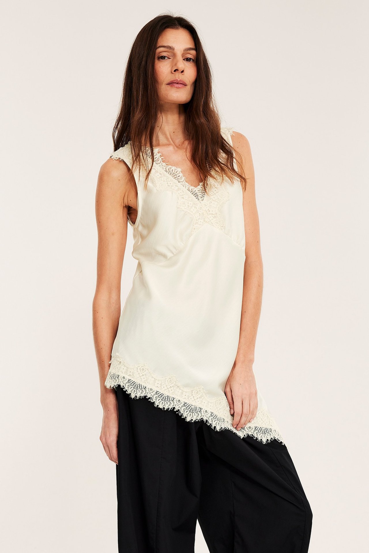 Kobieta nosi ecru satynowy top z koronkowym dekoltem w serek, asymetrycznym dołem, na czarnych spodniach.