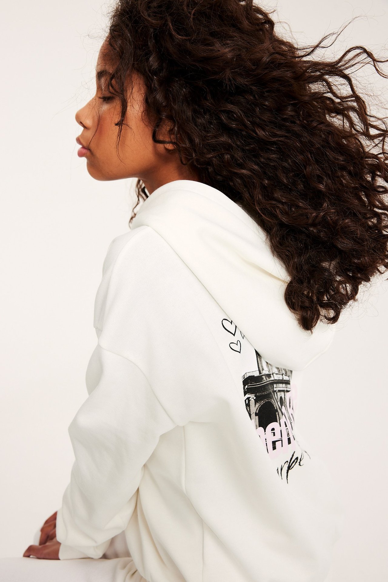 Widok z boku dziecka w ecru bawełnianej bluzie z kapturem z czarno-różowym nadrukiem na plecach.